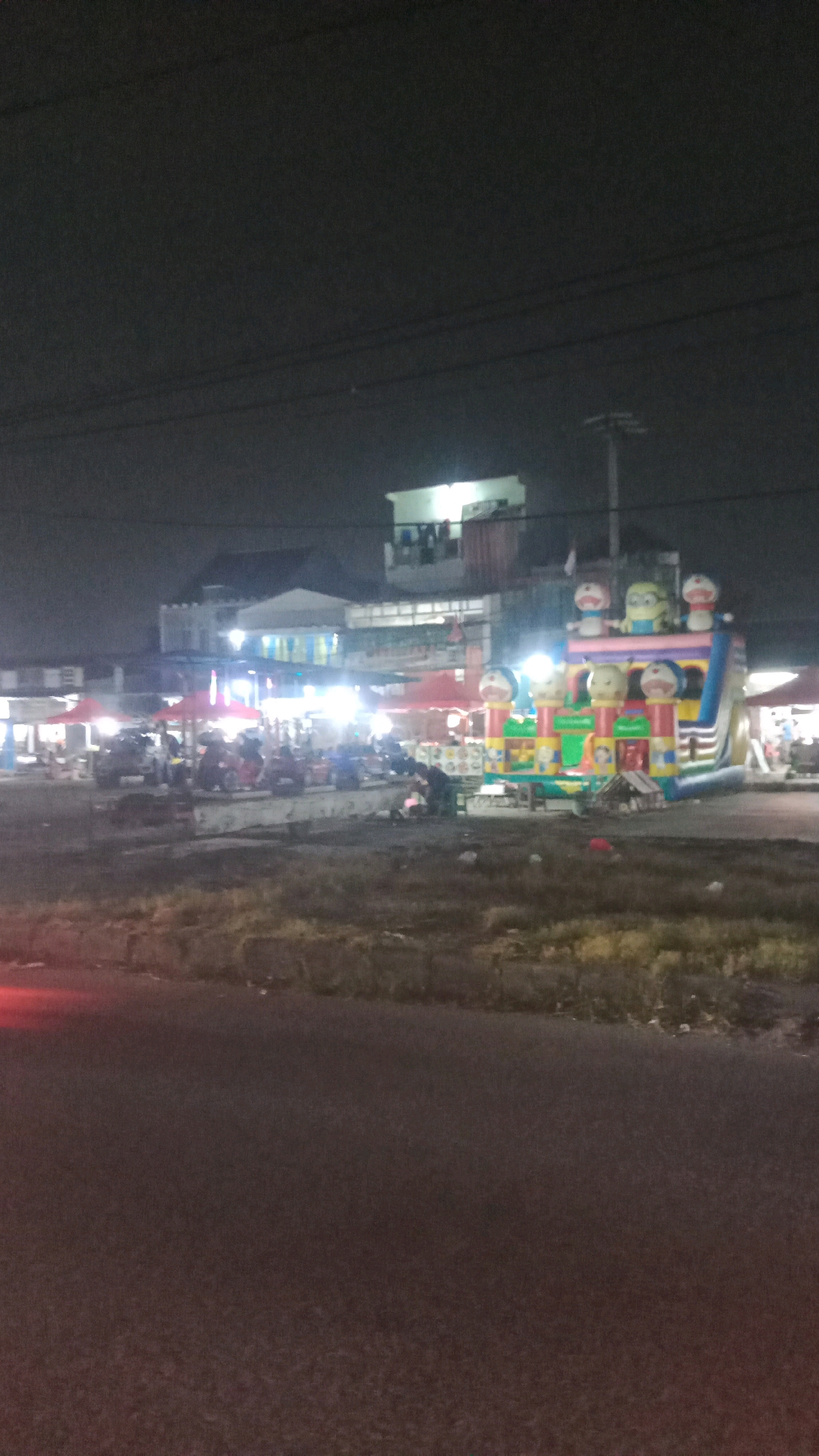 The image depicts an outdoor scene at night, likely in Jalenjaya, Indonesia. A prominent inflatable bouncy castle, decorated with cartoon characters resembling Doraemon and Minions, dominates the right side of the frame. It is illuminated and appears to be a point of attraction. In the background, there are multi-story buildings with brightly lit windows, suggesting commercial or residential establishments. Various vehicles, including motorcycles and what appear to be food stalls with red umbrellas, are visible in the middle ground, indicating a bustling marketplace or entertainment area. Several figures, appearing to be people, are dispersed among the stalls and around the bouncy castle, though their activities are indistinct due to the low light and image resolution. The foreground is a paved road, with a grassy verge bordering it. The overall impression is of a lively evening scene, possibly a local festival or market.