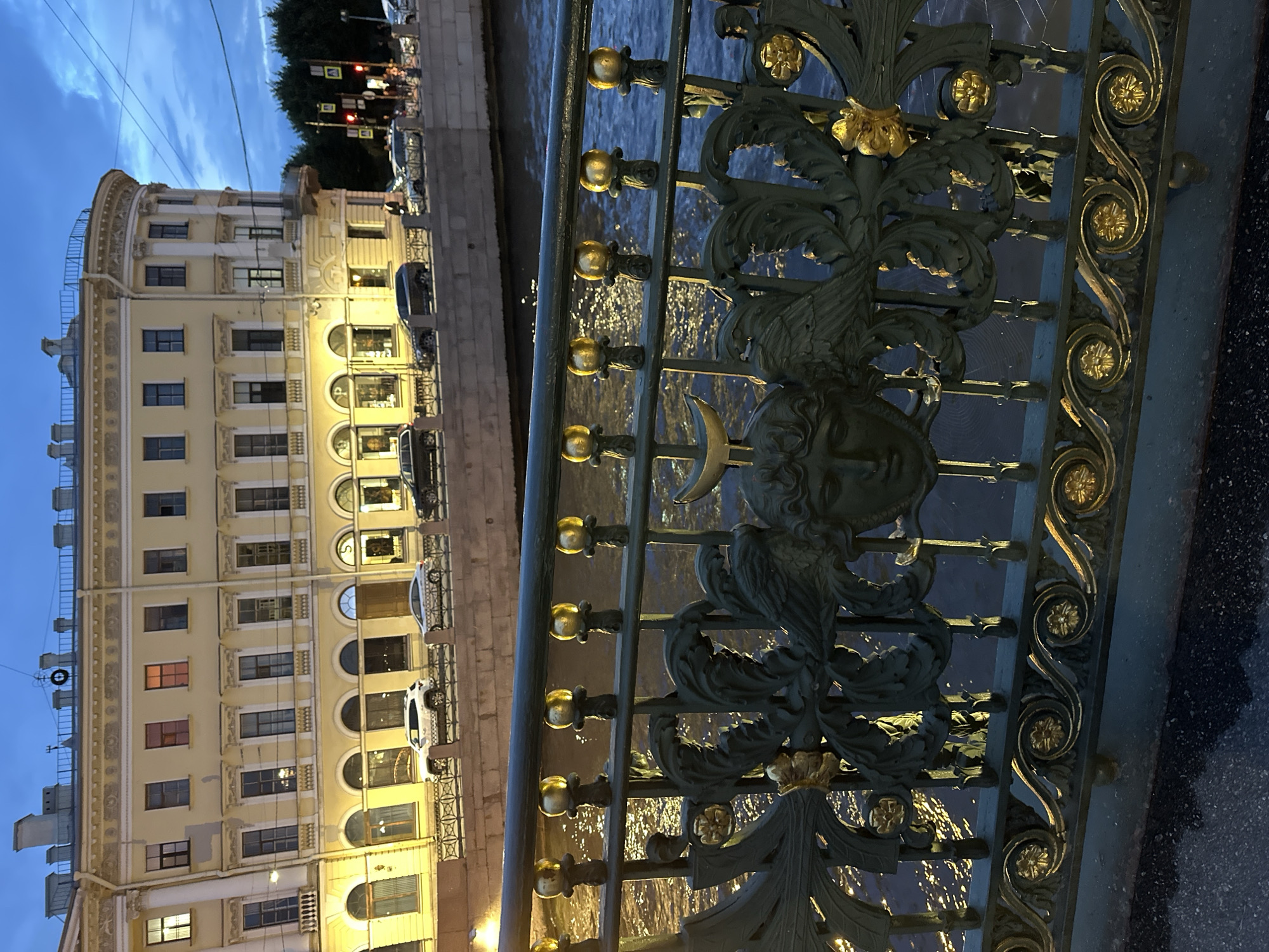 The image is a medium shot of a decorative metal railing in front of a building and a canal at dusk in Saint Petersburg, Russia. The railing is dark green with gold accents and features ornate designs, including faces and floral motifs. Behind the railing, a canal with illuminated water flows, reflecting the lights of the city. Across the canal, a cream-colored building with many windows stands. The sky is a deep blue with some clouds, indicating the evening hour. The scene appears to be a picturesque, historic location within Saint Petersburg, likely a bridge or embankment overlooking a canal.