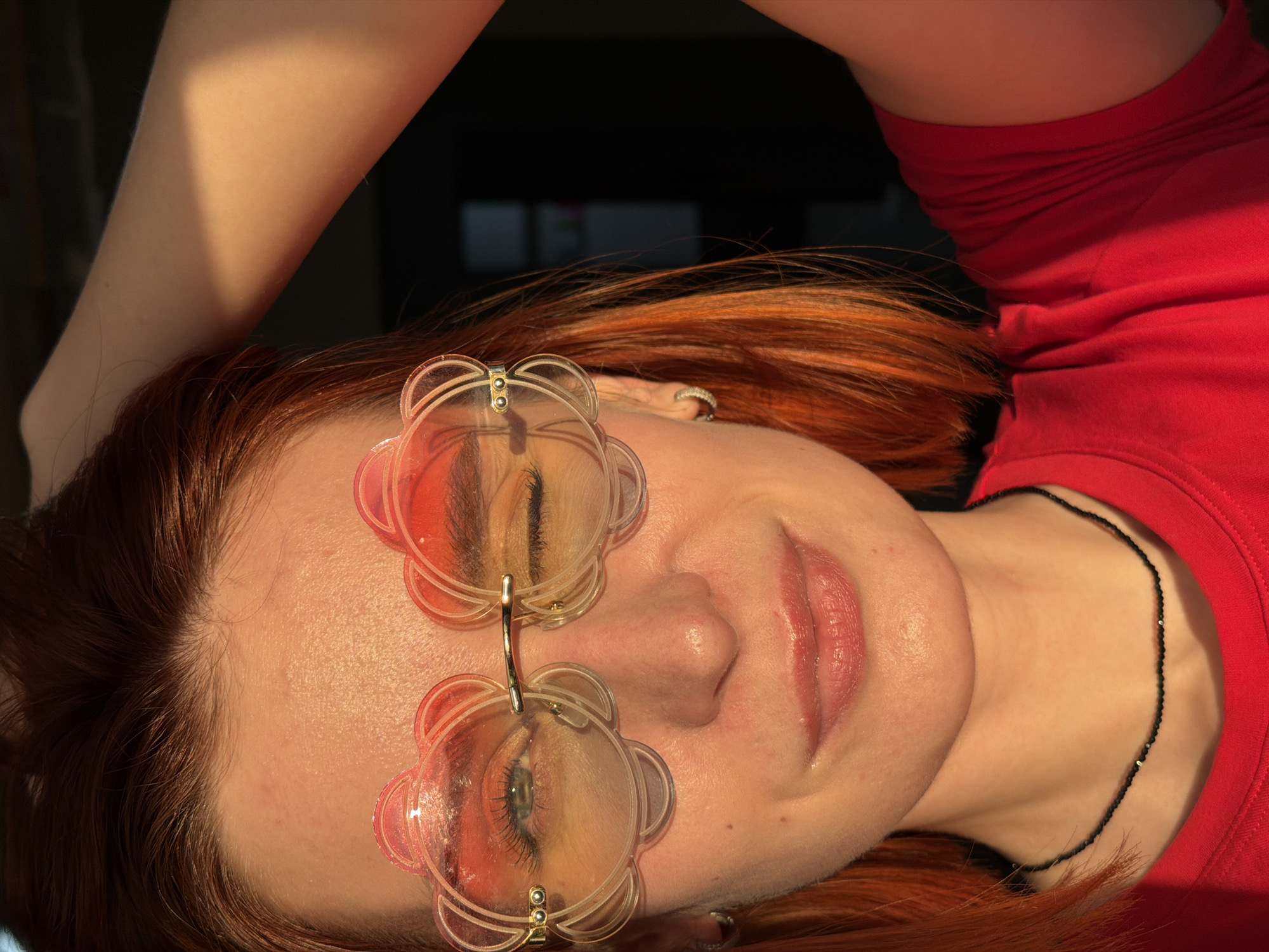 A close-up portrait features a female individual with reddish-orange, shoulder-length hair and light-toned skin, strongly illuminated by warm, directional light from the upper left. She is wearing clear, rimless sunglasses with two overlapping, stylized flower or cloud-shaped lenses per eye, exhibiting an orange-pink gradient tint. Her left eye (viewer's right) is open, revealing a blue-green iris, while her right eye (viewer's left) is closed in a wink. A subtle smile is present on her lips. She wears a small, silver-colored hoop earring on her left earlobe and a thin, black beaded necklace around her neck. Her right arm (viewer's left) is raised, with her forearm positioned above her head. She is dressed in a bright red crew-neck t-shirt. The background is dark and out of focus, suggesting an indoor or deeply shaded environment. The scene is located in the city of Chaikovsky, Russia.