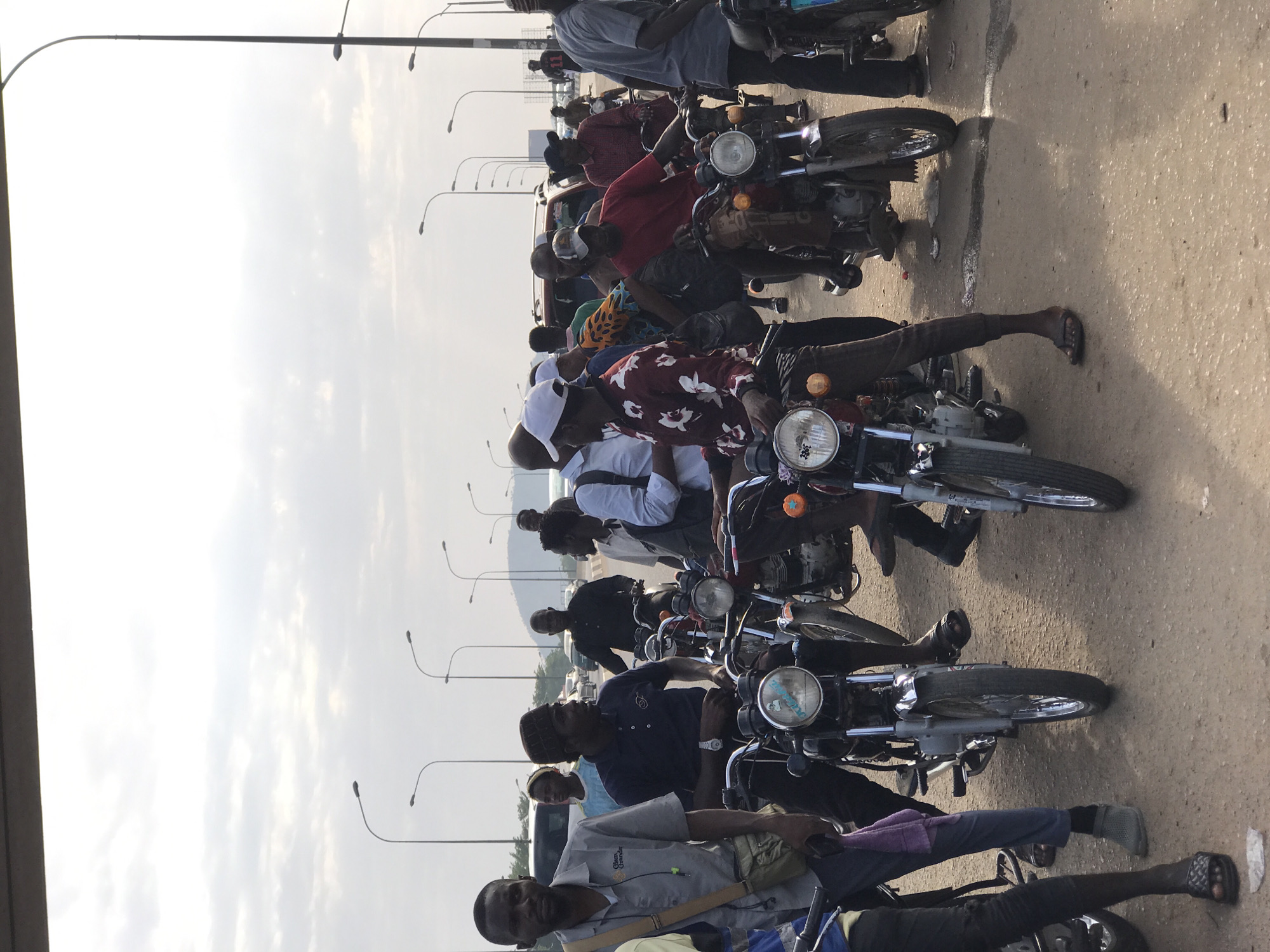 A medium shot shows a line of motorcycles parked on a paved road, likely a bridge or elevated roadway. Many riders are on their motorcycles. Riders are diverse in clothing. Street lights are visible in the background. The overcast sky suggests the image was taken in the daytime. Likely the location is Gwarinpa, Nigeria.