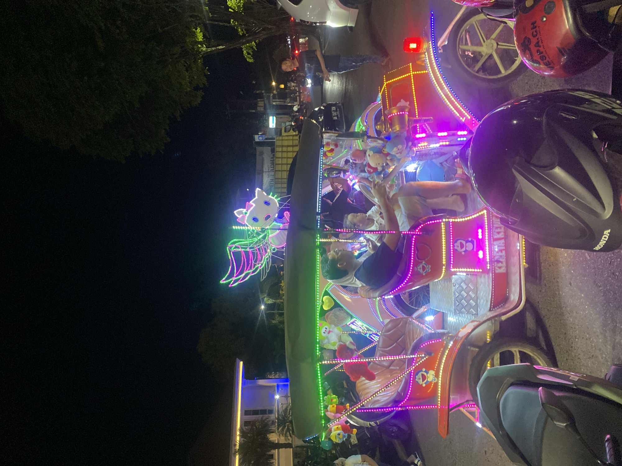 A neon-lit, open-air vehicle, resembling a decorated rickshaw, is parked on a paved street at night in Banjarbaru, Indonesia. The vehicle is adorned with numerous colorful LED lights in pink, green, and yellow, and features cartoon character decorations, including Hello Kitty and Doraemon. Several people are seated inside and on the vehicle, including a driver in the front and passengers in the back. The scene is illuminated by the vehicle's lights, streetlights, and ambient city lighting. In the foreground, two motorcycle helmets are visible, one black and one red. The background shows trees, buildings, and other vehicles, indicating an urban environment. The overall impression is one of a lively nighttime street scene, possibly a tourist attraction or a form of local transportation.