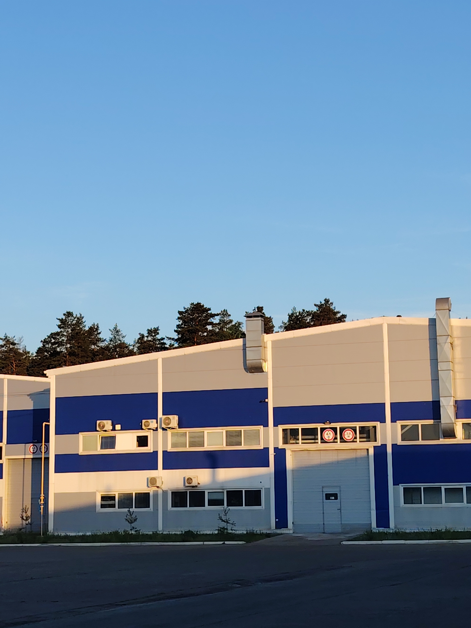 The image depicts a modern industrial building located in Noviy, Russia. The building's facade is characterized by alternating horizontal stripes of blue and gray, a common design element in modern industrial architecture. The building has a flat roof and is topped with several vertical ventilation or exhaust ducts. Along the facade are numerous windows, some of which are partially obscured by shadows, and several air conditioning units are mounted on the exterior walls. A large garage door occupies a central portion of the building, and signs displaying height restrictions are visible above the door. The presence of small trees planted in front of the building suggests a landscaped area. The building appears to be situated in an industrial or commercial zone, as evidenced by the paved area in the foreground. The sky is clear and blue, indicating fair weather conditions. The background features a line of trees, suggesting the building is located on the outskirts of the city or near a green space. There is no visible human activity, suggesting the scene is either unpopulated or was captured during a period of inactivity.