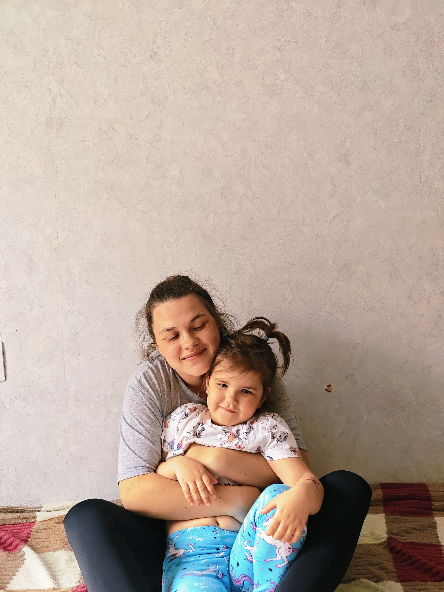 A young woman with her hair in a messy bun is sitting cross-legged, holding a young girl in her lap. The woman is wearing a gray t-shirt and dark leggings. The girl is wearing a white shirt with unicorn designs and blue leggings with unicorn designs. They are both looking at the camera and smiling slightly. They are sitting on a patterned blanket. The background is a plain gray wall. The image was taken indoors, likely in a private residence. The location is Chaikovsky, Russia.