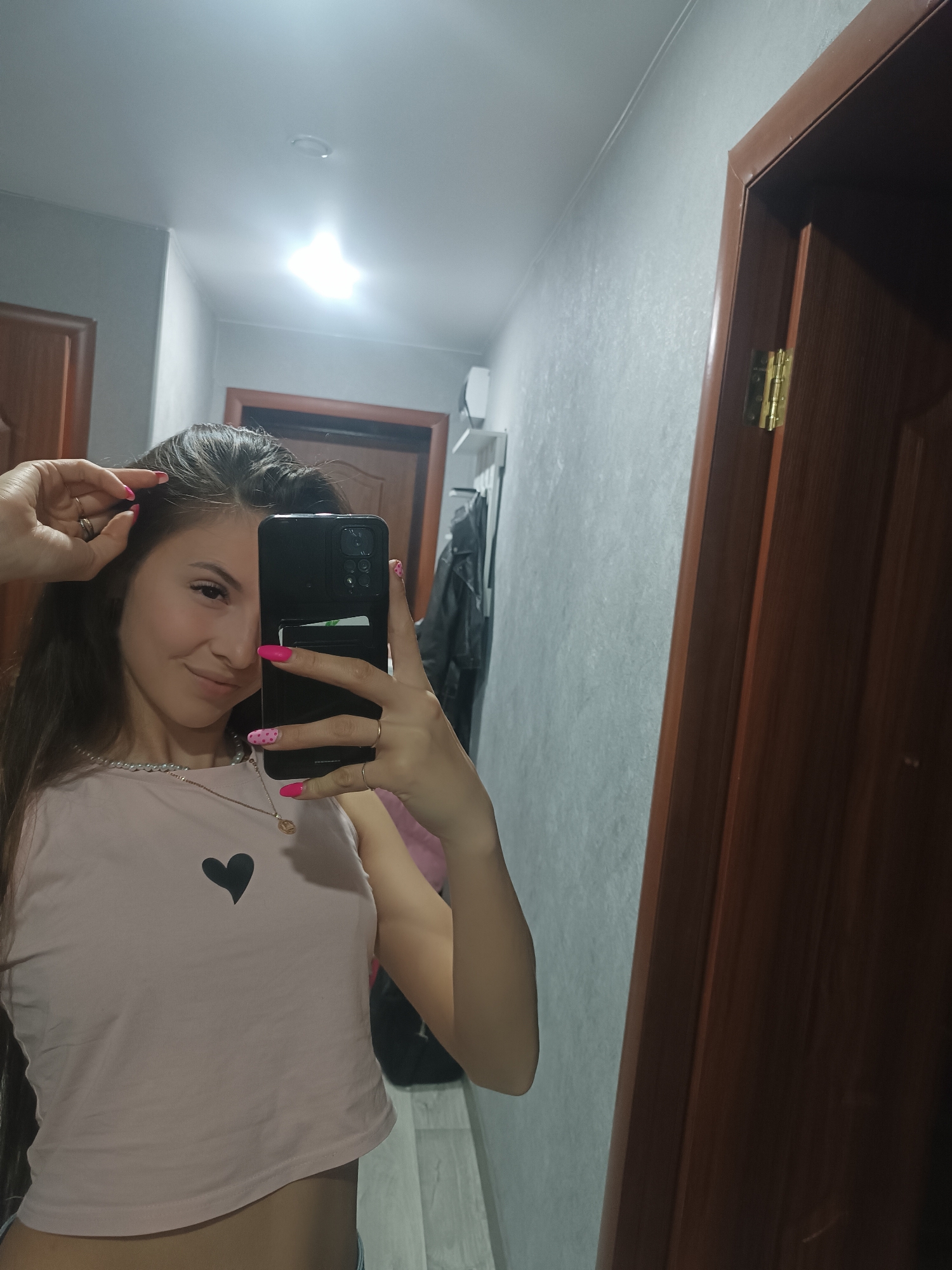A person is taking a selfie in what appears to be a hallway. The person is holding a black smartphone in front of their face, partially obscuring it. They are wearing a light pink crop top with a black heart on the front. The person has long dark hair. The location of the hallway in the city of Unknown, Unknown is not discernible from the provided image.