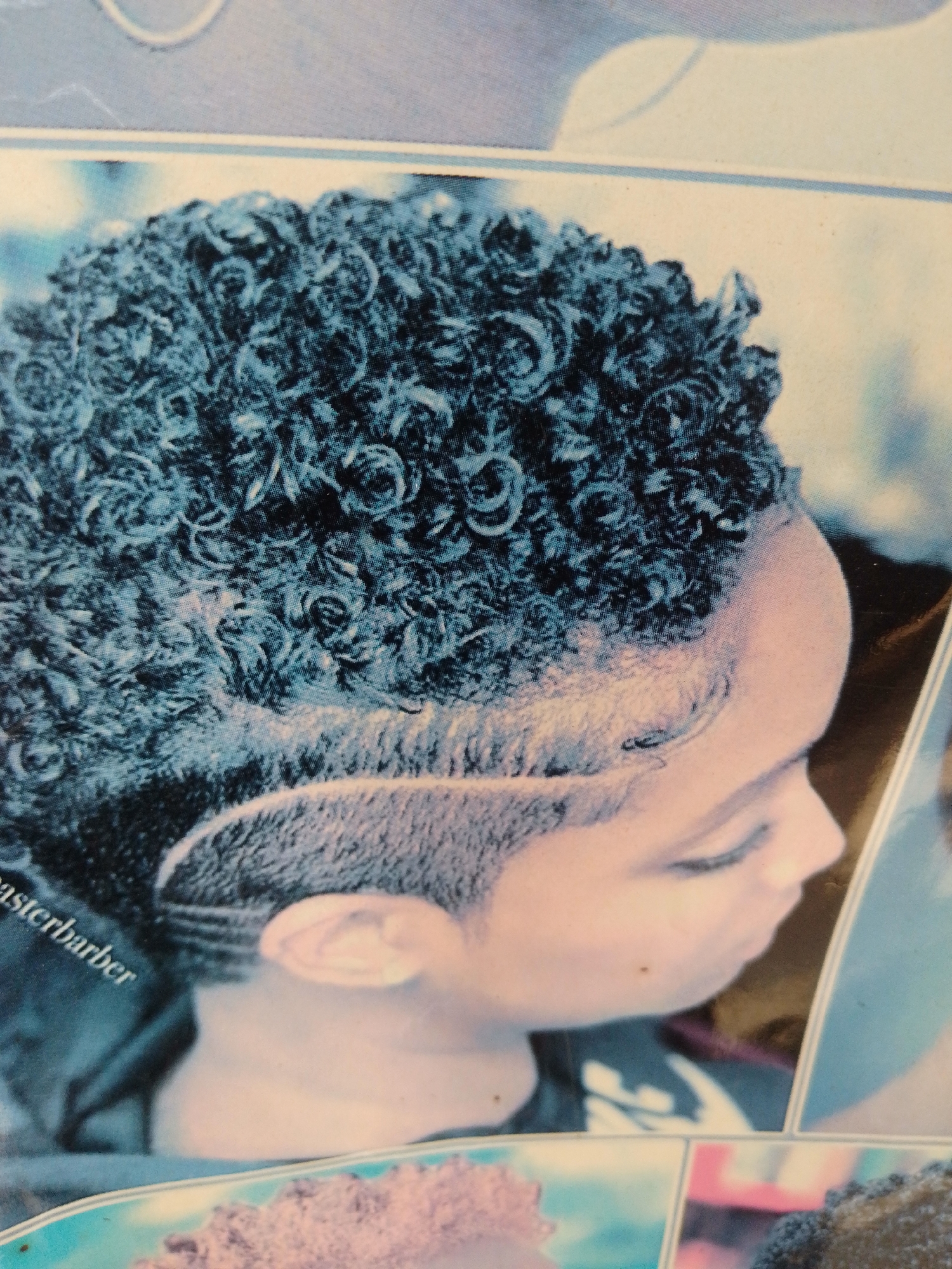 The image is a close-up, high-angle shot of a young Black person's head from the side, with their hair styled in tight, dark curls. The person's skin is light brown. A sharp, carved line runs along the side of their head, creating a distinct contrast with the shorter, faded hair below. The background is blurred and features abstract shapes and colors, suggesting it might be a photograph from a magazine or a display. The person's eye is visible and appears to be closed or looking down. The overall lighting is cool, with blue and white tones dominating the image. The text "masterbarber" is visible on the left side of the image.