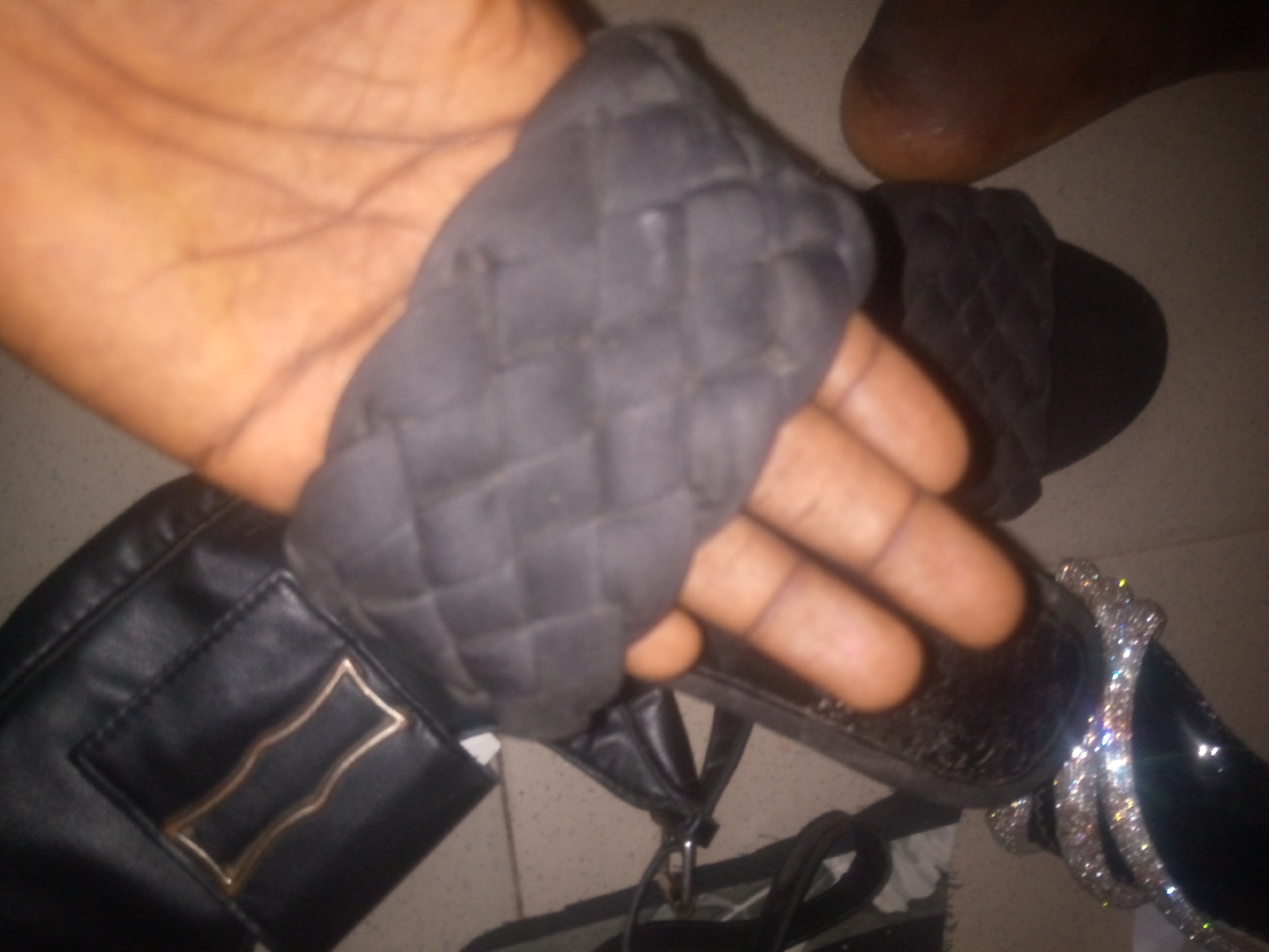A close-up, overhead shot shows a person's hand holding a black, quilted sandal. The sandal has a woven strap and is resting on another black shoe. The background is blurry but appears to be a tiled floor. The scene is set in a room in Jalingo, Nigeria.