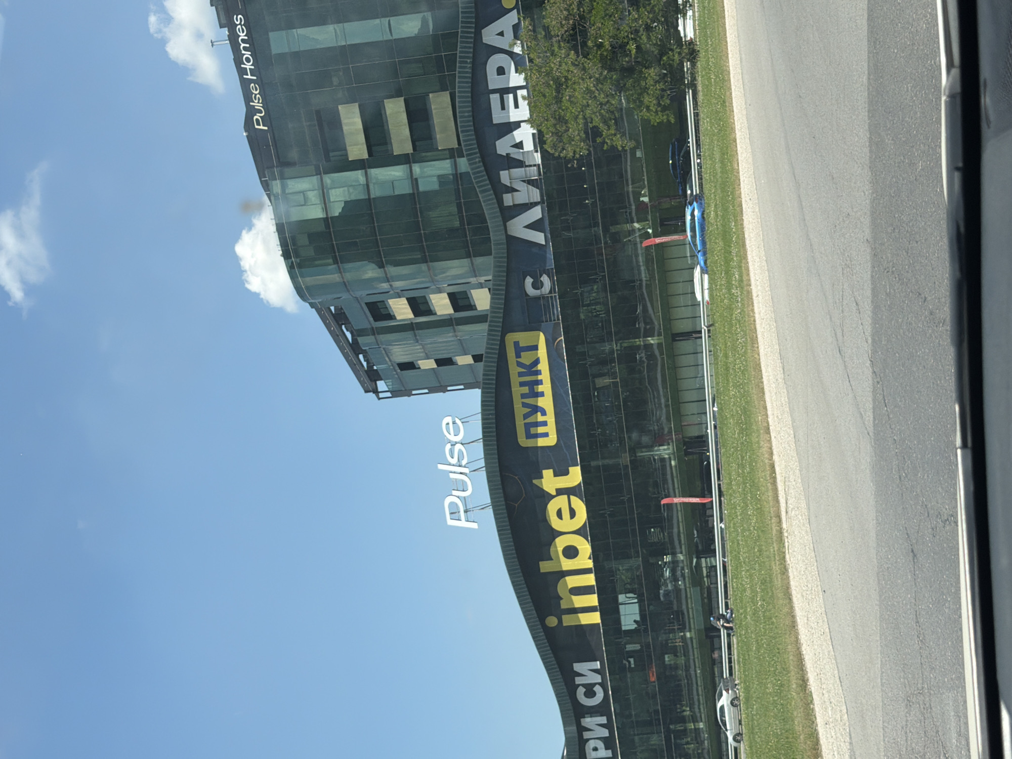 A modern, multi-story building with a curved, reflective glass facade stands under a bright blue sky with scattered white clouds. The building displays prominent signage. On the lower section, a large banner features yellow lettering spelling "inbet" and "ПУНКТ" within a yellow rectangle, alongside Cyrillic text. Above this, a white sign reads "Pulse." Higher up on the building's dark roof, white lettering states "Pulse Homes." The building is situated in a semi-urban environment, with a grassy median strip in the foreground bordering a road. Several cars are visible on the road and the median, including a blue station wagon and a silver sedan. A person on a scooter is also present on the grassy median. The overall scene suggests a commercial or business district.