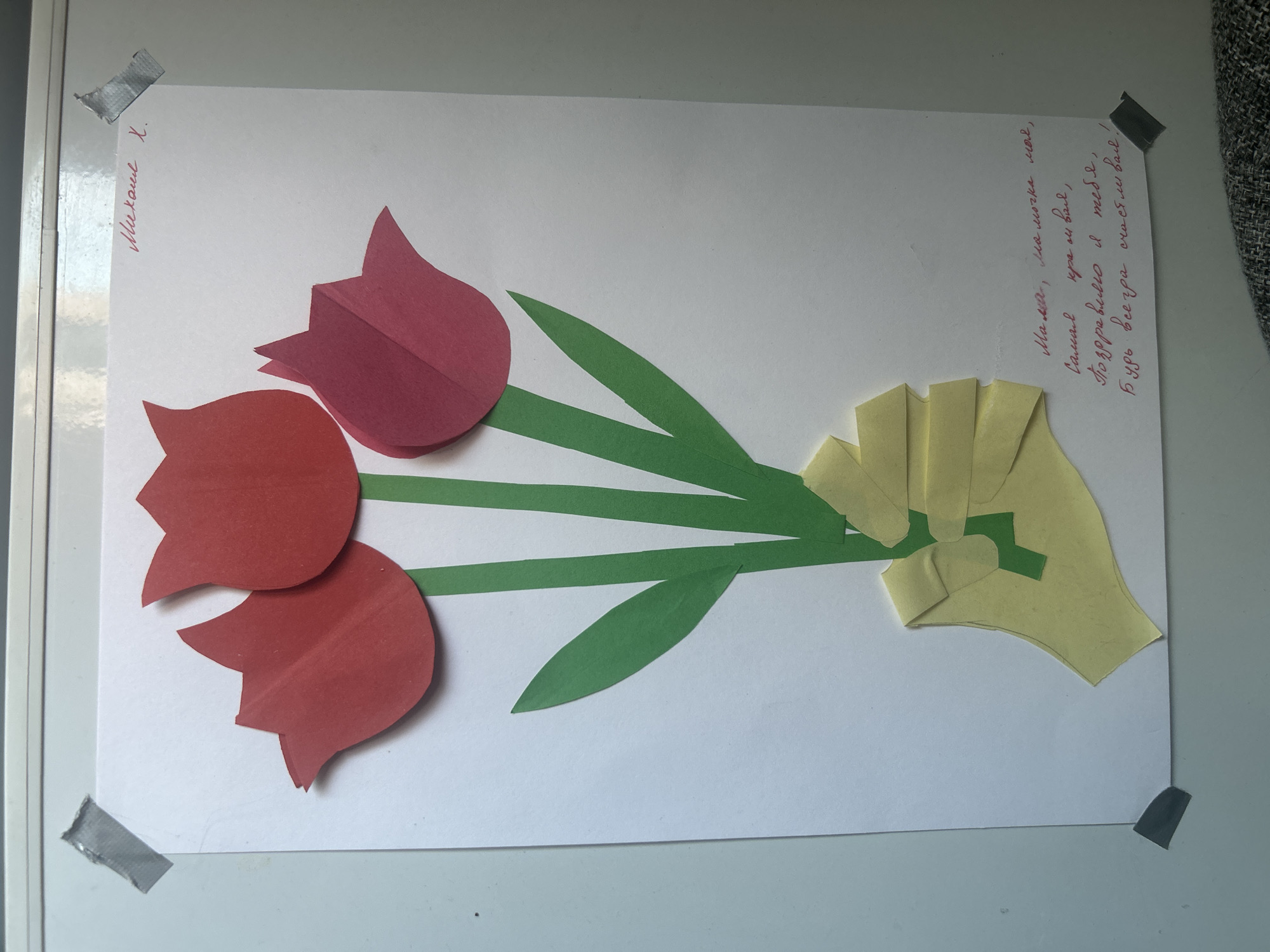 The image displays a piece of paper with a collage of paper tulips and a hand holding them. The tulips are made from red and pink paper, and their stems and leaves are made from green paper. The hand is made from yellow paper. Below the tulips, there is handwritten text in Russian. The paper is attached to a white surface with grey tape in the corners. There are no visible people or indications of the city of Kant, Kyrgyzstan. The scene is a close-up of a craft project.