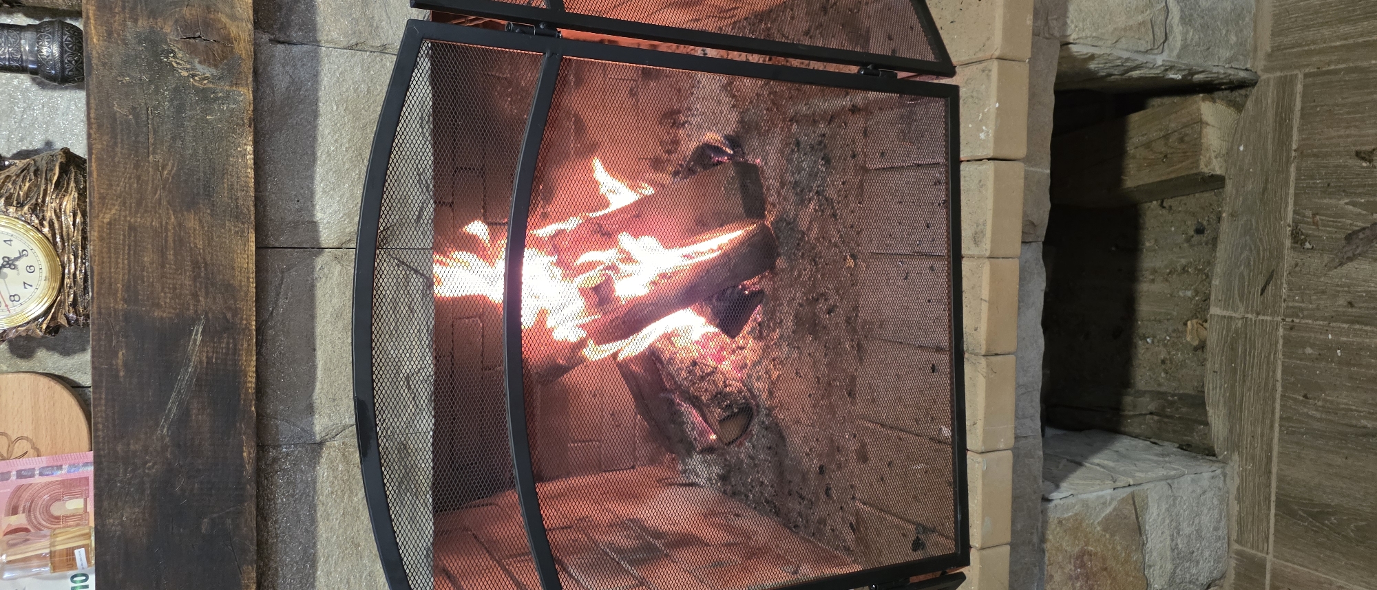 The image shows a fireplace with a protective screen in Novachene, Bulgaria. Burning logs are visible behind the screen. To the left of the fireplace, against a stone wall, there is a decorative wall clock, a wooden cutting board and some euro banknotes. To the right of the fireplace is what appears to be a niche in the wall. The construction material looks to be bricks.