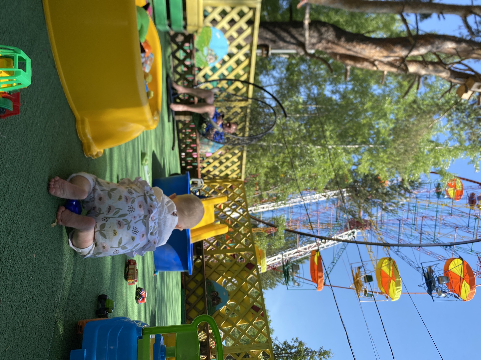 The image captures a scene in Chaikovsky, Russia, featuring a child on green artificial turf in an outdoor play area. The child, wearing a patterned romper, is on their hands and knees. The area includes yellow and blue plastic play structures. A person is sitting in a swing suspended from a tree. In the background is a Ferris wheel against a clear blue sky.