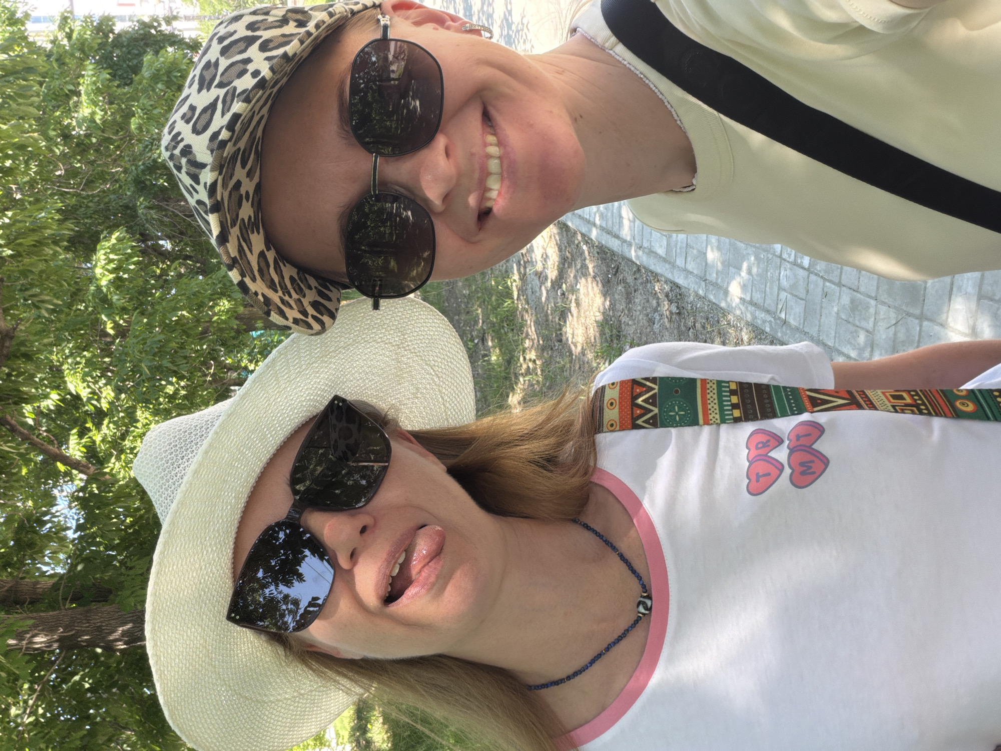 The image shows two women taking a selfie outdoors, possibly on a sunny day in a park or along a sidewalk. The woman on the left is wearing a wide-brimmed white hat, large dark sunglasses, and a white t-shirt with a pink collar. She has her tongue sticking out playfully. Around her neck is a beaded necklace. A colorful patterned strap is visible on her shoulder. The woman on the right is wearing a leopard-print baseball cap, dark sunglasses, and a light yellow t-shirt. She is smiling broadly. A black strap from a bag is visible across her body. The background shows trees with green foliage, suggesting a natural setting. The ground appears to be a paved walkway with a light grey brick pattern. Without further context or identifiable landmarks, it is not possible to definitively place this image in the city of Khabarovsk, Russia. The image captures a moment of casual interaction between two individuals enjoying an outdoor environment.
