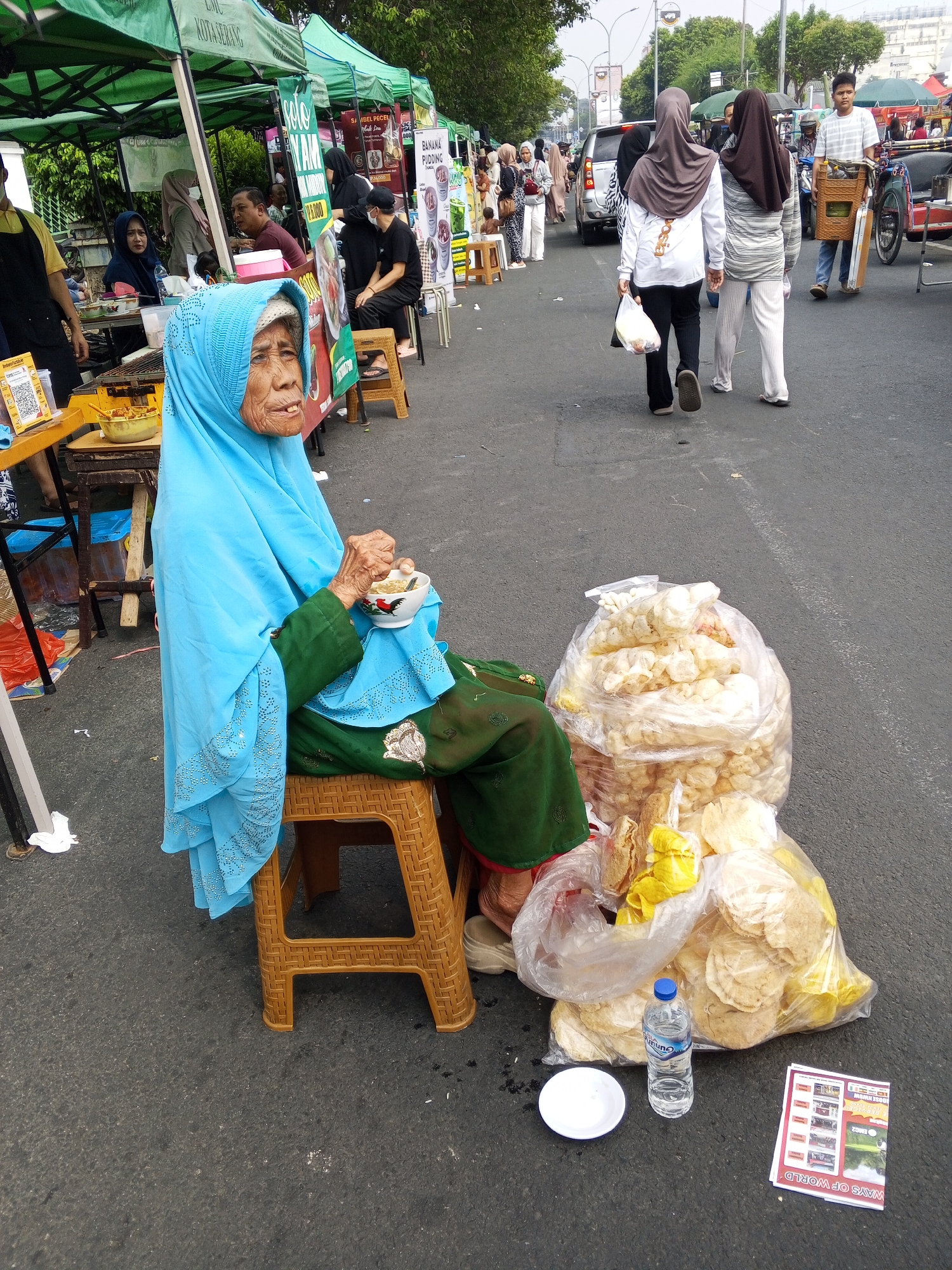 An elderly woman wearing a blue hijab and a green dress sits on a plastic stool at an outdoor market in Serang, Indonesia. She holds a small bowl of food in her hands, appearing to be eating. Several large plastic bags filled with what appear to be crackers or snacks are placed around her. In front of her, on the asphalt ground, is a white bowl, a bottle of water, and a pamphlet. In the background, there are market stalls with green awnings, other people browsing, and vehicles on the street, suggesting a bustling urban environment. The overall scene captures a moment of daily life at a roadside market.