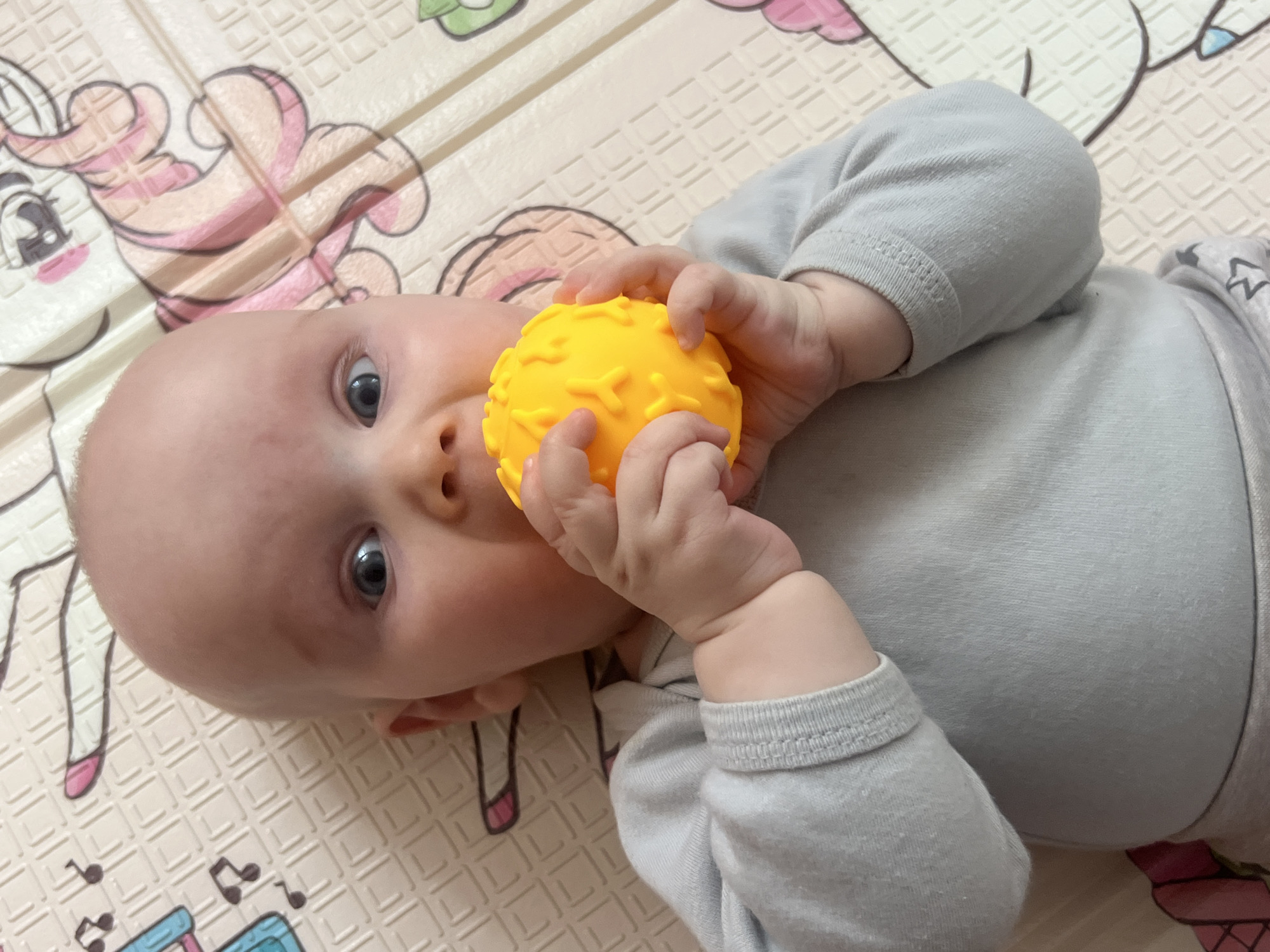 A baby is lying on a play mat, facing the camera at an angle. The baby is light-skinned, with a bald head and blue eyes. They are wearing a gray long-sleeved shirt and holding a yellow textured ball with both hands. The ball is positioned near the baby's mouth. The play mat has a light beige background with cartoon illustrations. No information is available about a location in Votkinsk, Russia.