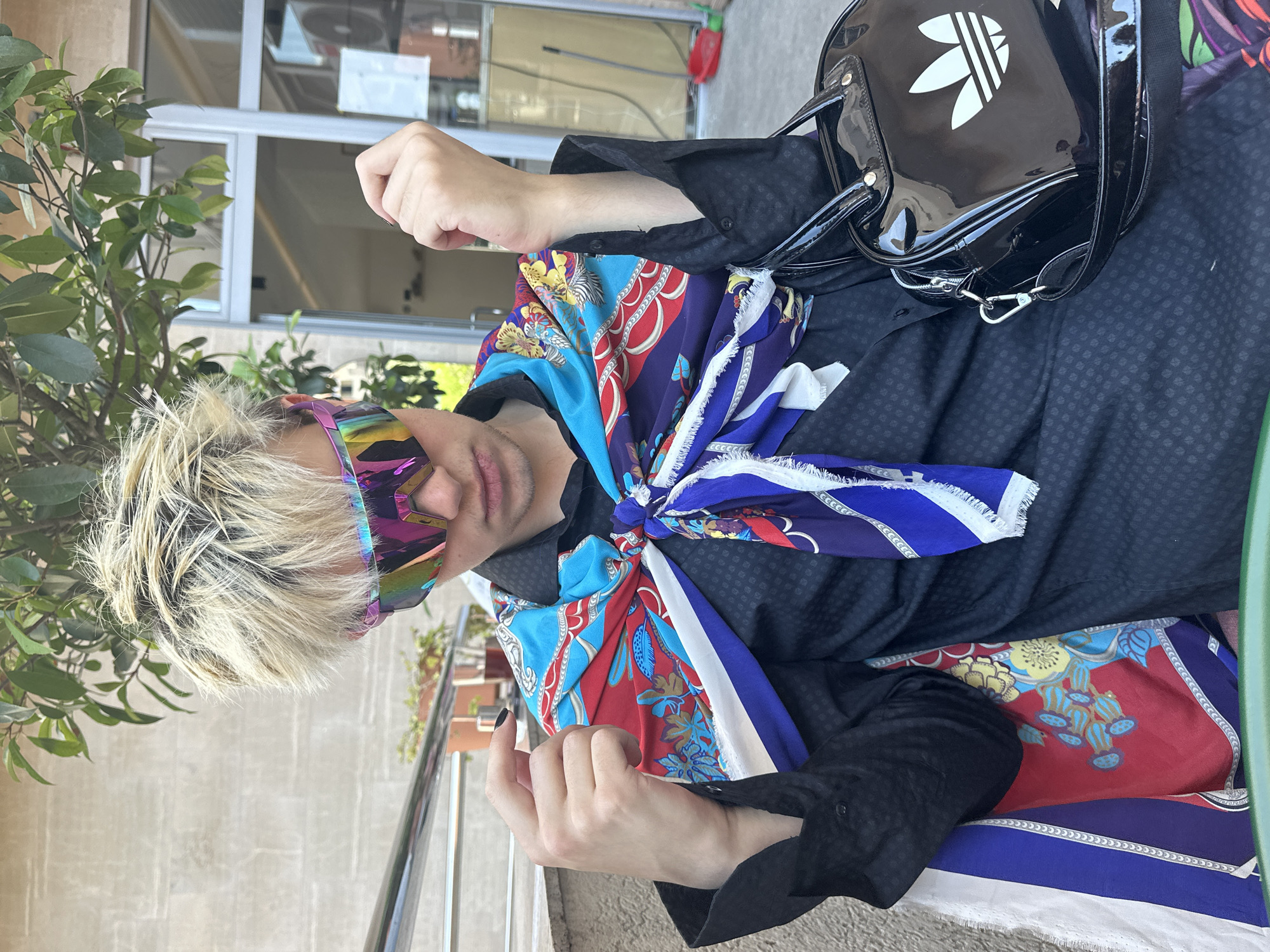 A person with bleached, spiky hair wearing oversized, iridescent sunglasses is seated outdoors. They are dressed in a black button-down shirt adorned with a colorful, patterned scarf tied around their neck and draped over their shoulders. A shiny black handbag with a white Adidas logo is placed on their right side. The background shows a stone wall, some foliage, and a doorway with glass panels. The scene appears to be in an outdoor seating area, possibly a cafe or restaurant, located in Budva, Montenegro.