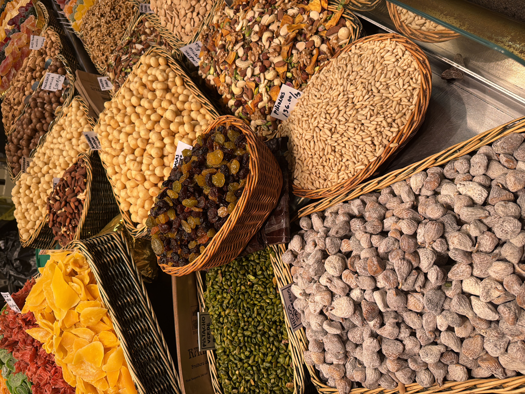 The image shows a variety of dried fruits and nuts displayed in wicker baskets. There are several types of nuts and dried fruits are present including pistachios, macadamia nuts, raisins, and figs. Labels are visible with text and pricing information. Text on one of the labels indicates the location is "Mcat. Boqueria 540 541", which refers to a stall located in the Boqueria Market in Barcelona, Spain.