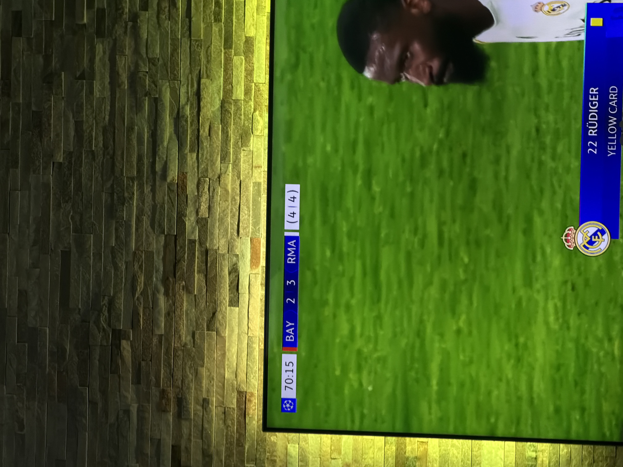 The image shows a television screen displaying a soccer game, likely a Champions League match based on the branding and logo. The game's score is visible, indicating BAY 2 - 3 RMA with a time of 70:15. A yellow card has been issued to player 22 Rüdiger. The TV screen is mounted on a wall covered with stacked stone veneer panels. No individuals or other discernible elements are present in the vicinity of the wall. There is no indication this picture was taken in the city of Buhovo, Bulgaria.