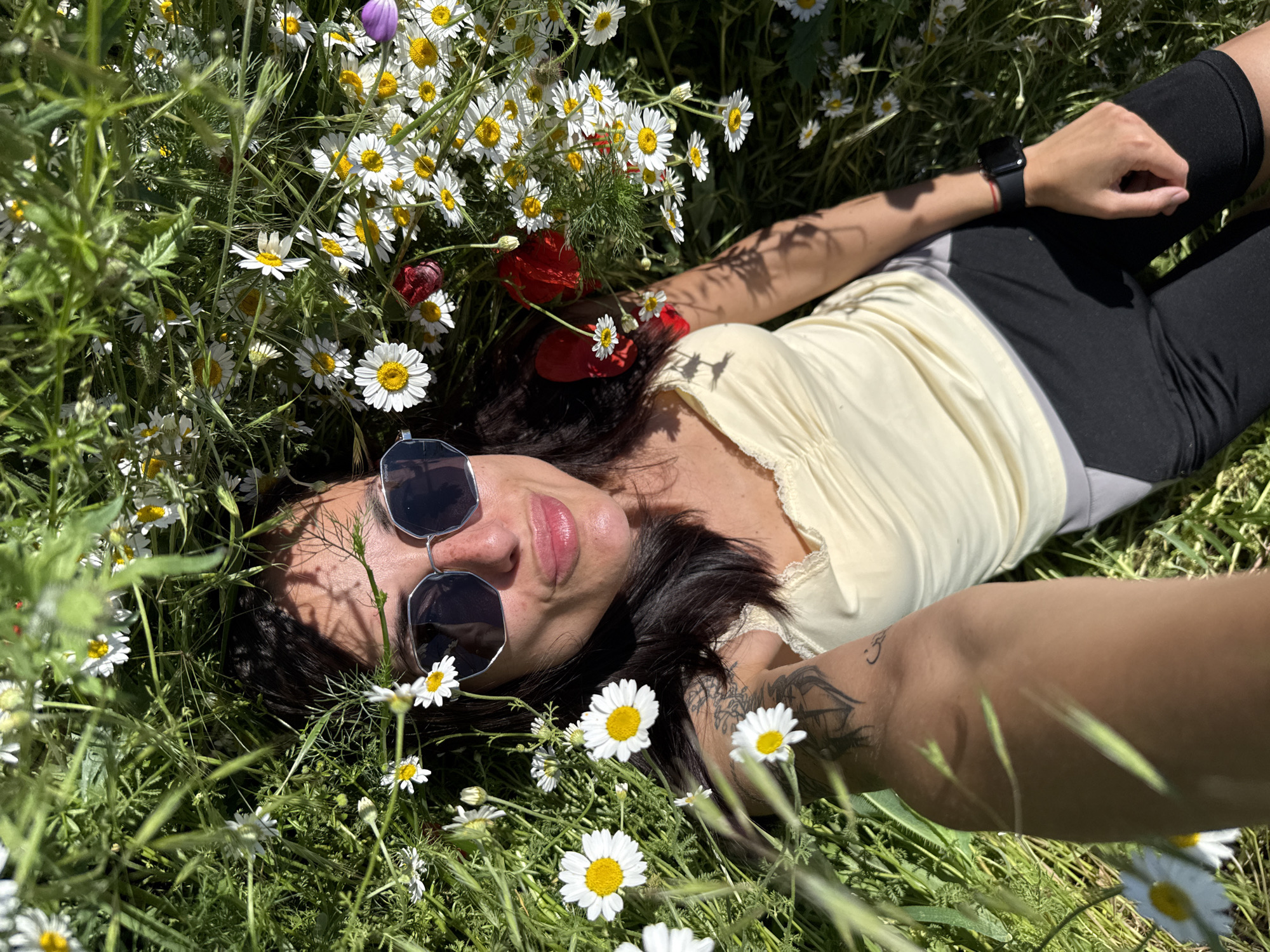A woman lies in a field of daisies and grass. She is wearing sunglasses, a pale yellow top, and black shorts. A smartwatch is visible on her wrist. She appears to be taking a selfie, as her arm is extended towards the camera. The scene suggests a sunny, outdoor location with abundant wildflowers. I cannot provide the precise location in Pleven, Bulgaria without further information.