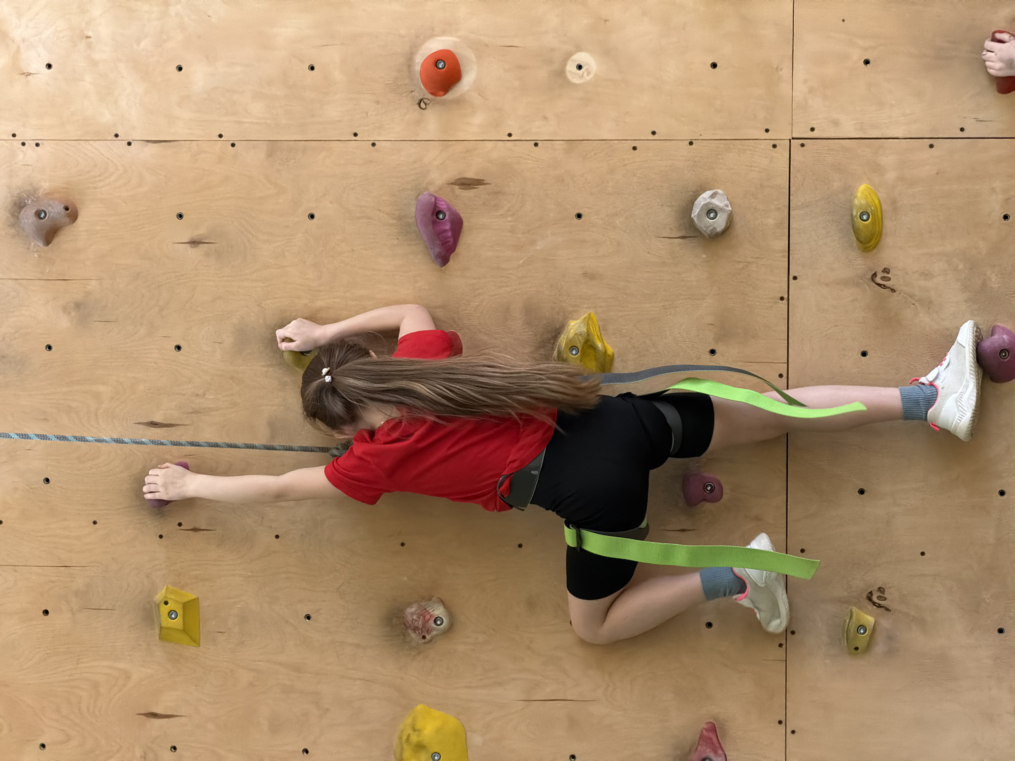 An overhead shot captures a young person, likely a girl, engaged in climbing or traversing a wooden indoor climbing wall in Izhevsk, Russia. The climber is stretched horizontally across the frame, wearing a red t-shirt, black shorts, and white sneakers with gray socks. A black climbing harness with bright green webbing is worn around their waist and legs, and a blue and white speckled climbing rope runs horizontally along the wall, connected to the harness, providing safety or assistance.
The climber's long, brown hair is tied back with white ties. Their right hand is extended, reaching for a purple climbing hold, while their left hand grasps another purple hold. Both feet are placed on various colorful climbing holds (purple, yellow, and red). The climbing wall consists of multiple light-colored wooden panels, secured with numerous small black bolts, and features a variety of textured and brightly colored holds in shades of orange, purple, yellow, red, and light gray. The scene depicts a focused and active moment within an indoor climbing facility. There is no visible text in the image.