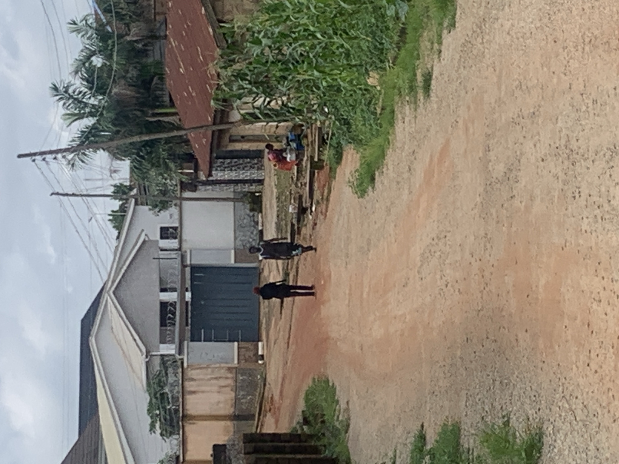 The image depicts a dirt road in a residential area, likely in Jos, Nigeria. Two individuals, one in a dark shirt and trousers and the other in a dark t-shirt and shorts, walk away from the camera down the road. On the right side of the road, a woman in a red and pink outfit is seated near a building with a corrugated iron roof, tending to a basin and some containers. Lush green vegetation, including corn stalks, grows next to the building. Across the road, a white building with a large, dark blue gate is visible behind a beige wall and fence. Palm trees and utility poles with electrical wires are present in the background, suggesting an urban or semi-urban environment. The sky is overcast with clouds.