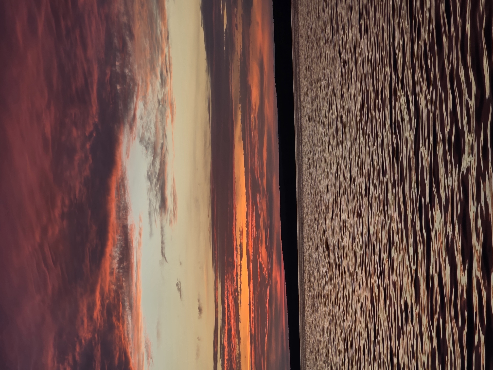 The image captures a vibrant sunset over a calm body of water, likely the Kama River in Chaikovsky, Russia. The sky is a dramatic canvas of deep reds, oranges, and purples, with wispy clouds reflecting the fiery hues of the setting sun. A dark, silhouetted treeline forms the horizon, separating the sky from the rippling water. The water's surface mirrors the sunset colors, creating a textured pattern of light and shadow. No people or man-made structures are visible, emphasizing the natural beauty of the scene.