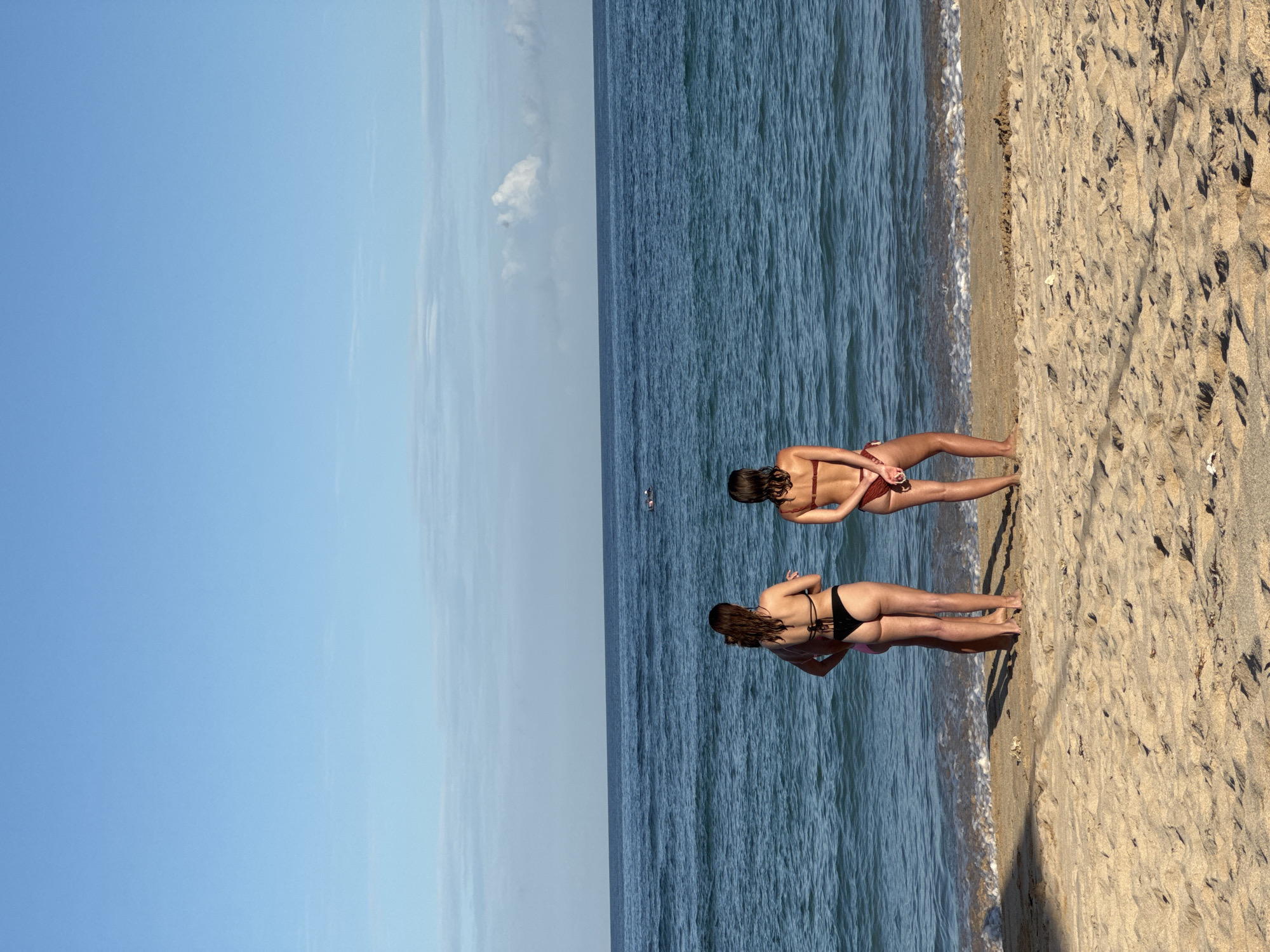 The image depicts a beach scene in Kuta Selatan, Indonesia. Two women in bikinis are standing on the sand at the edge of the ocean. The sky is blue with some light clouds. The ocean water is a deep blue color. The sand is light brown and appears to be dry.