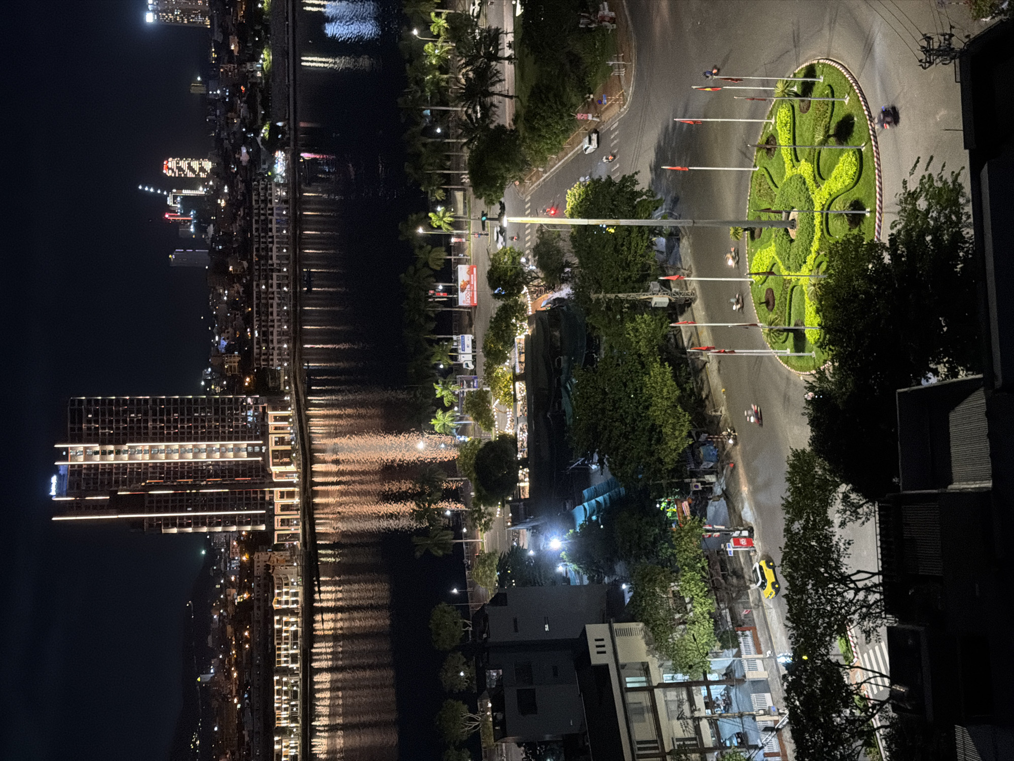 This is a night aerial view of a coastal city, likely Đà Nẵng, Vietnam, featuring a modern cityscape along a waterfront. In the foreground, a roundabout with a circular, green landscaped island is visible, adorned with flags on poles. Several cars and motorcycles are depicted on the surrounding roads, with some in motion, indicated by motion blur. Palm trees line the waterfront and urban areas.
The midground showcases urban development with buildings of varying heights, including a prominent, illuminated high-rise skyscraper with vertical lighting. The buildings are reflected in the dark water of a river or bay that separates the foreground from the distant cityscape. The reflection of lights from buildings and the sky creates a shimmering effect on the water's surface.
The background displays a broader view of the city's skyline, with numerous lights indicating urban activity. Distant hills or mountains are visible behind the city. The overall scene conveys a sense of a vibrant, developed urban environment at night, with a focus on the interplay of light, architecture, and natural elements like the water and vegetation.