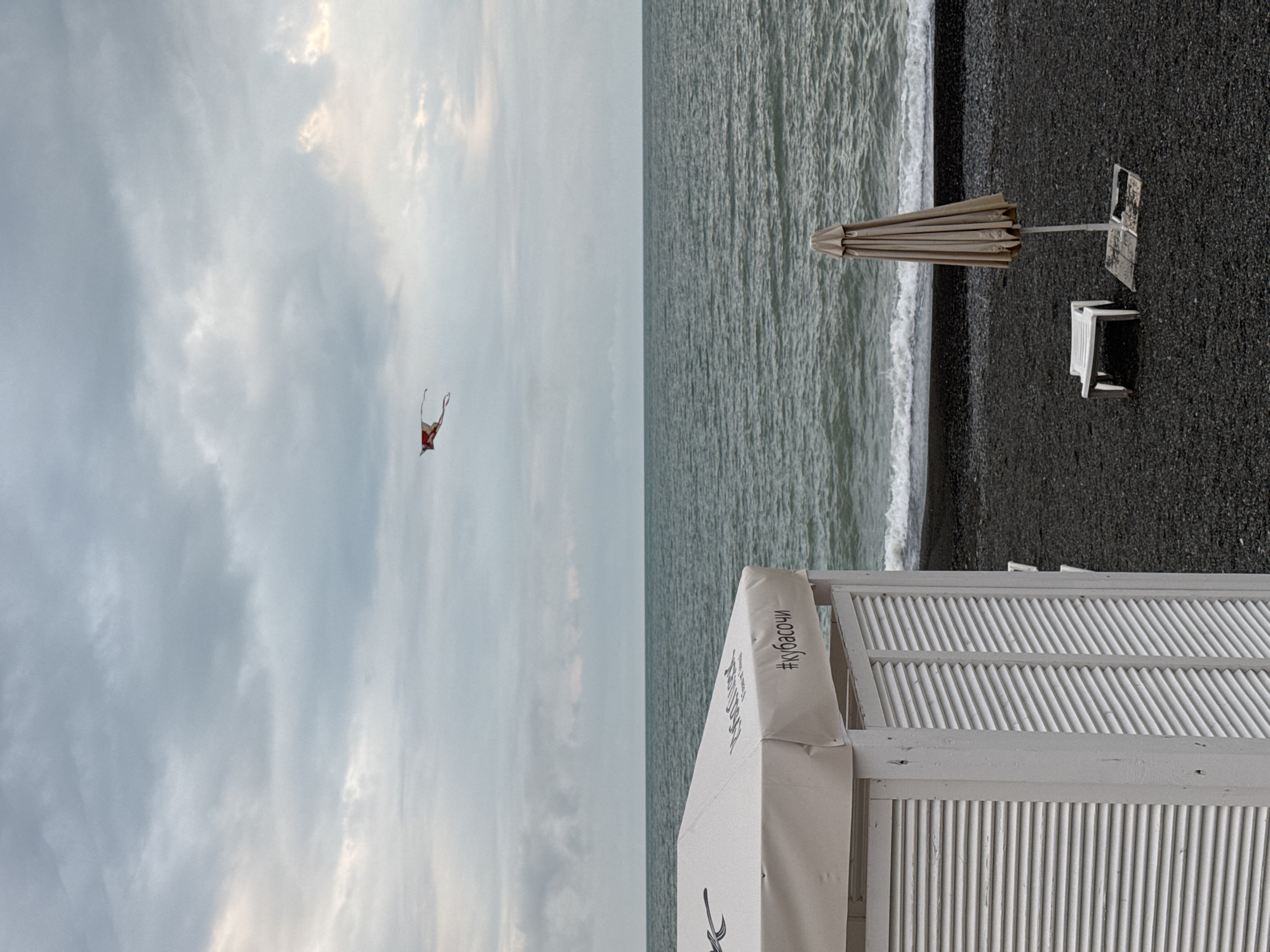 A vertically-oriented photograph depicts a beach scene, likely located in Sochi, Russia, indicated by the "#кубасочи" tag on a structure. The beach is composed of dark gravel or sand and is bordered by the ocean. An umbrella and two chairs are on the beach. A white, louvered structure stands in the foreground. A kite is in the sky. The sky is overcast.