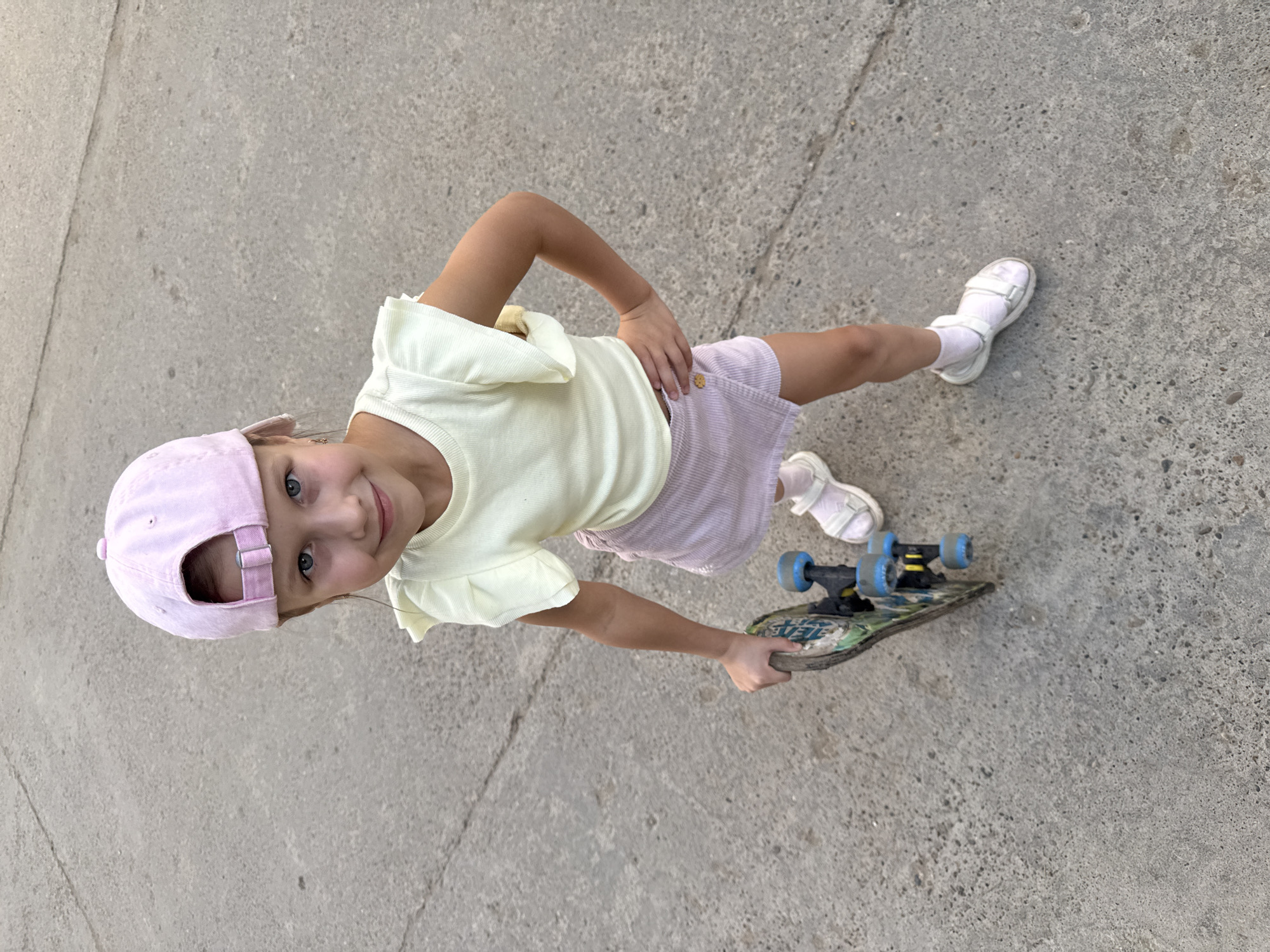 A young girl stands on a concrete surface, likely a sidewalk or paved area. She wears a light yellow top with frilled sleeves, a striped pink skirt, white socks and sandals, and a pink baseball cap. She holds a skateboard with blue wheels in her right hand. Her left hand rests on her hip. She is looking up at the camera. The location is likely Chaikovsky, Russia based on the prompt, but there are no features visible to confirm this.