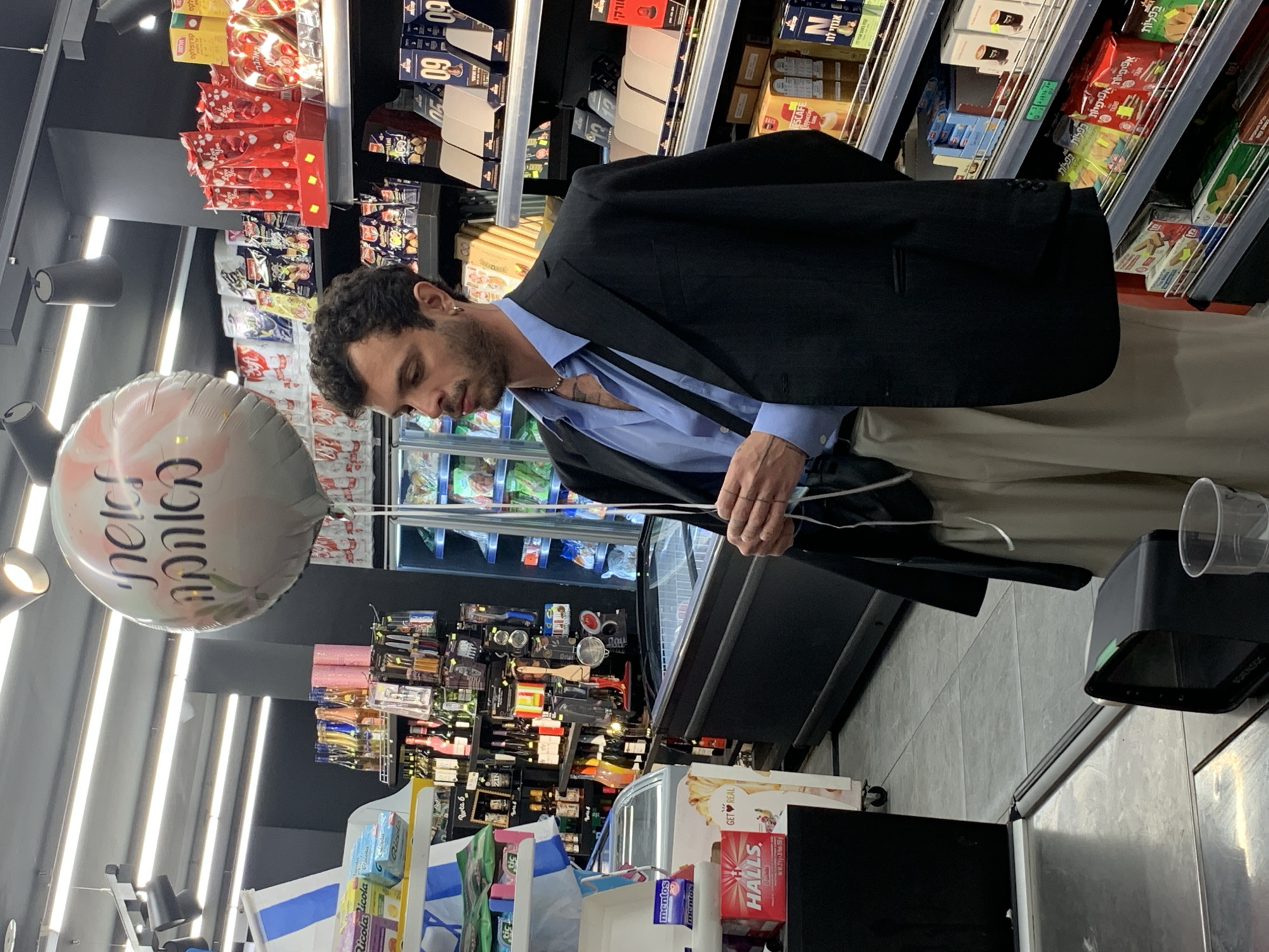 The scene is an indoor setting, likely a supermarket or grocery store, in Tel Aviv, Israel, given the Hebrew lettering on products and a balloon. A man is at the checkout counter. He has dark curly hair and a beard, wearing a light blue button-down shirt, a black blazer draped over his shoulder, and beige pants. He is holding the strings of a helium balloon with Hebrew text on it, standing next to a refrigerated display case.
Various food products line the shelves in the background. The foreground features the checkout counter with a cash register or monitor, plastic cups, and other retail items. Packages with familiar brands like Ricola, Mentos, Halls, and Nescafé are visible.