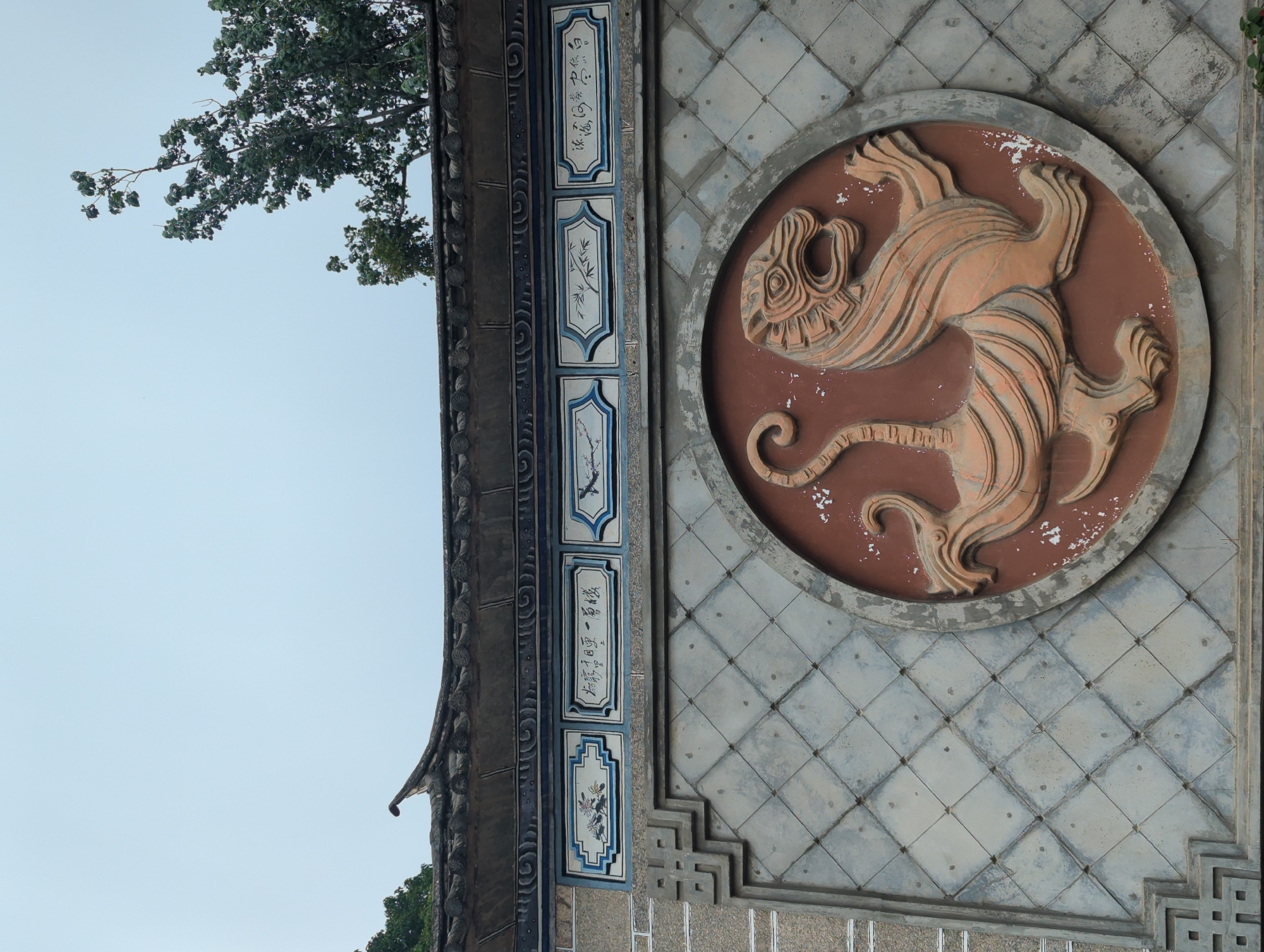This image shows a close-up of an ornate wall in Xishan District, China, featuring a bas-relief sculpture of a tiger within a circular frame. The tiger is depicted in a dynamic pose, with its mouth open and mane flowing. The wall itself is constructed of grey, diamond-patterned tiles and adorned with blue and white decorative panels above the tiger sculpture. These panels contain Chinese calligraphy and floral or botanical motifs. The roofline of a traditional Chinese building with curved tiles is visible at the top of the image, along with a portion of a tree. The sky is overcast.