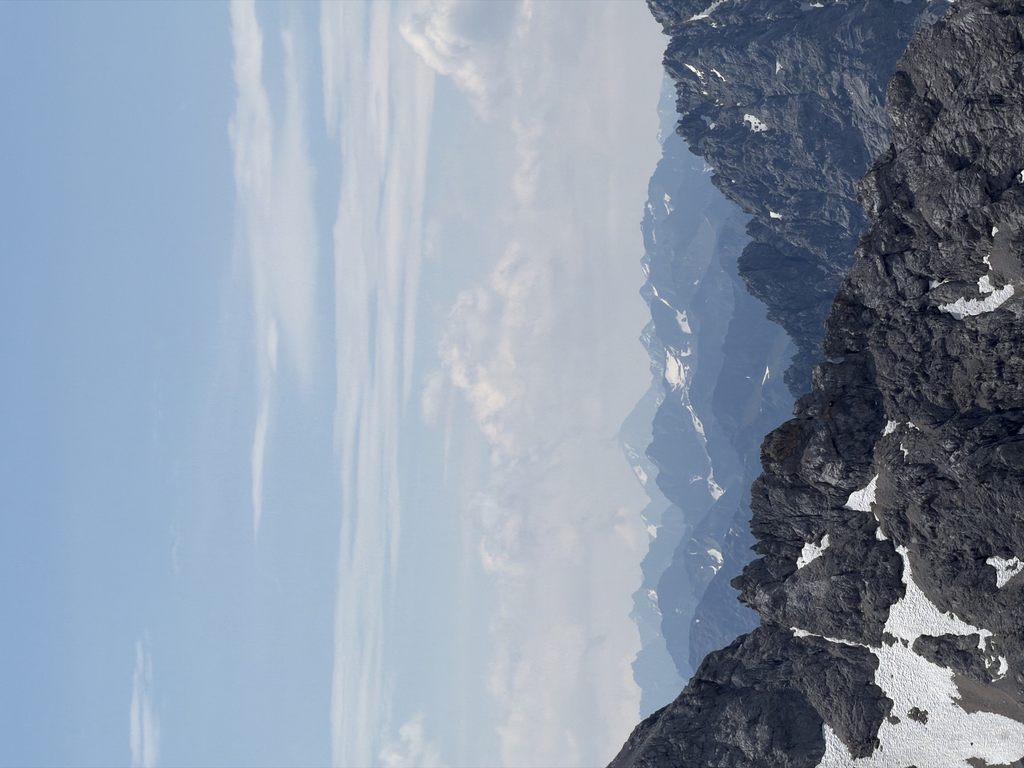 This media file is a photograph of a mountainous landscape, likely taken from a high vantage point. The scene is dominated by rugged, dark grey rock formations with patches of snow clinging to their surfaces. These foreground mountains are sharply defined and textured, suggesting a rocky and uneven terrain.
Behind the immediate peaks, a series of receding mountain ranges are visible, becoming progressively hazier and lighter in color, creating a sense of depth and distance. These distant mountains also show signs of snow cover, particularly in the shaded valleys and on the higher slopes. The sky is a clear, pale blue, with wispy white clouds stretching across it. The clouds are thin and elongated, possibly indicating high-altitude cirrus clouds.
The lighting suggests it is daytime, likely late morning or early afternoon, given the clear sky and the way the light illuminates the mountain faces. There are no people or objects visible in the image, focusing solely on the natural environment. The overall mood is one of grandeur and remoteness, highlighting the stark beauty of the alpine environment. The presence of snow even in what appears to be a clear sky suggests a high altitude location where snow can persist. Garmisch-Partenkirchen, Germany, is known for its dramatic alpine scenery, consistent with the image's depiction. No text is visible in the media file.