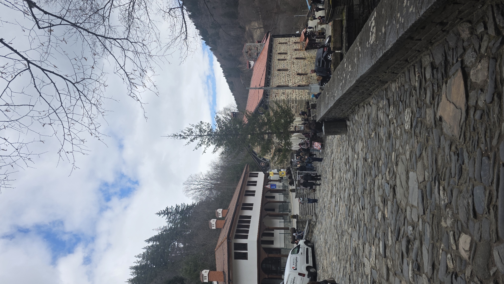 The scene depicts an outdoor area in Rilski manastir, Bulgaria, under a partially cloudy sky. In the foreground, a dark grey stone cobblestone pavement occupies the lower half of the image, leading towards a gathering of people. To the right, a low, dark stone retaining wall, approximately 0.75 meters high, runs parallel to the pavement.
Two prominent buildings are visible. On the left, a two-story building features white plastered walls, a red-tiled roof with multiple white chimneys, and a covered porch area with dark wooden columns and railings on the ground floor. Dark, rectangular windows are present on both floors. A white "InterLogistica" commercial van is parked near this building on the left.
To the right of the first building and slightly behind a tall, slender coniferous tree, stands a building constructed of varied grey stone blocks with a red-tiled roof. This building has arched, recessed windows on its side visible in the image. A "Shop" sign is discernible near its entrance on the far right.
Numerous individuals are present in the midground, forming several small groups and milling about. Some are walking, others appear to be conversing, and a few are seated at outdoor tables near the stone building on the right, which seems to host a market or cafe area with barrels and stalls. A dark SUV is parked near the stone building on the right.
The background features densely wooded hillsides covered with coniferous and leafless deciduous trees, indicating a mountainous or forested environment. Bare tree branches are visible at the top and right edges of the frame. A blue directional sign with a white arrow pointing left is visible near the white building.