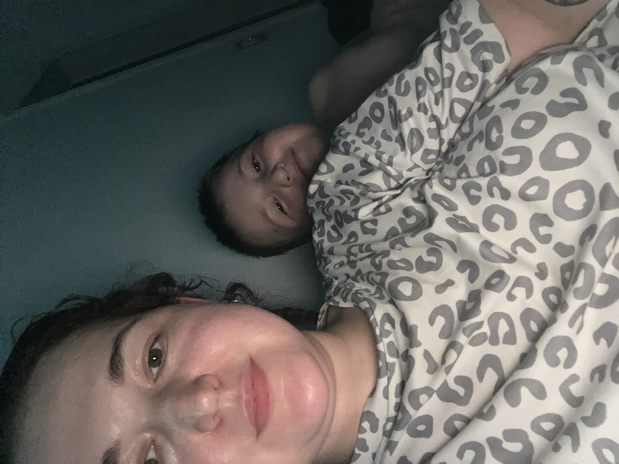 This is a close-up, low-light selfie of two individuals, likely a mother and child, in a domestic setting. The person in the foreground, to the left, is a woman with curly dark hair and fair skin, wearing a grey and white leopard-print shirt. Her face is close to the camera, and she appears to be looking slightly off-camera. Behind her, to the right, is a child with fair skin and light brown hair, also wearing a similar leopard-print garment. The child is leaning against something, possibly a pillow or the woman, and is looking directly at the camera with a slight smile. The background is dark and indistinct, with some shadows suggesting an indoor environment. There are no discernible features that would indicate a specific location within Chelyabinsk, Russia.
