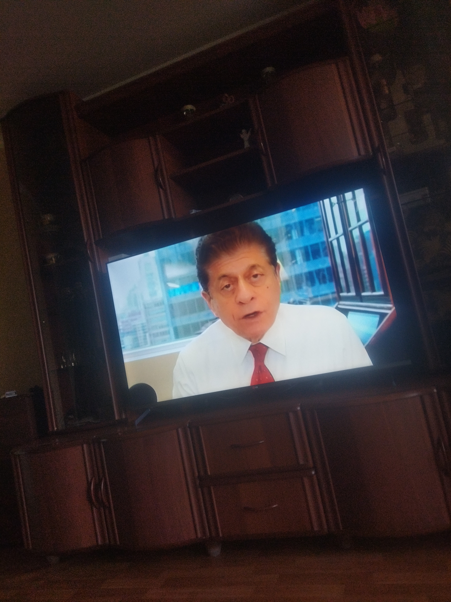 A television screen displays a man in a white shirt and red tie. He is looking to his left and speaking. Behind him, the screen shows a blurred cityscape with blue buildings and windows. The television is placed on a dark brown wooden entertainment center with multiple cabinets and shelves. The entertainment center is in a room with a dark wooden floor. The scene does not provide any information about the city of Qarağandı city administration, Kazakhstan.