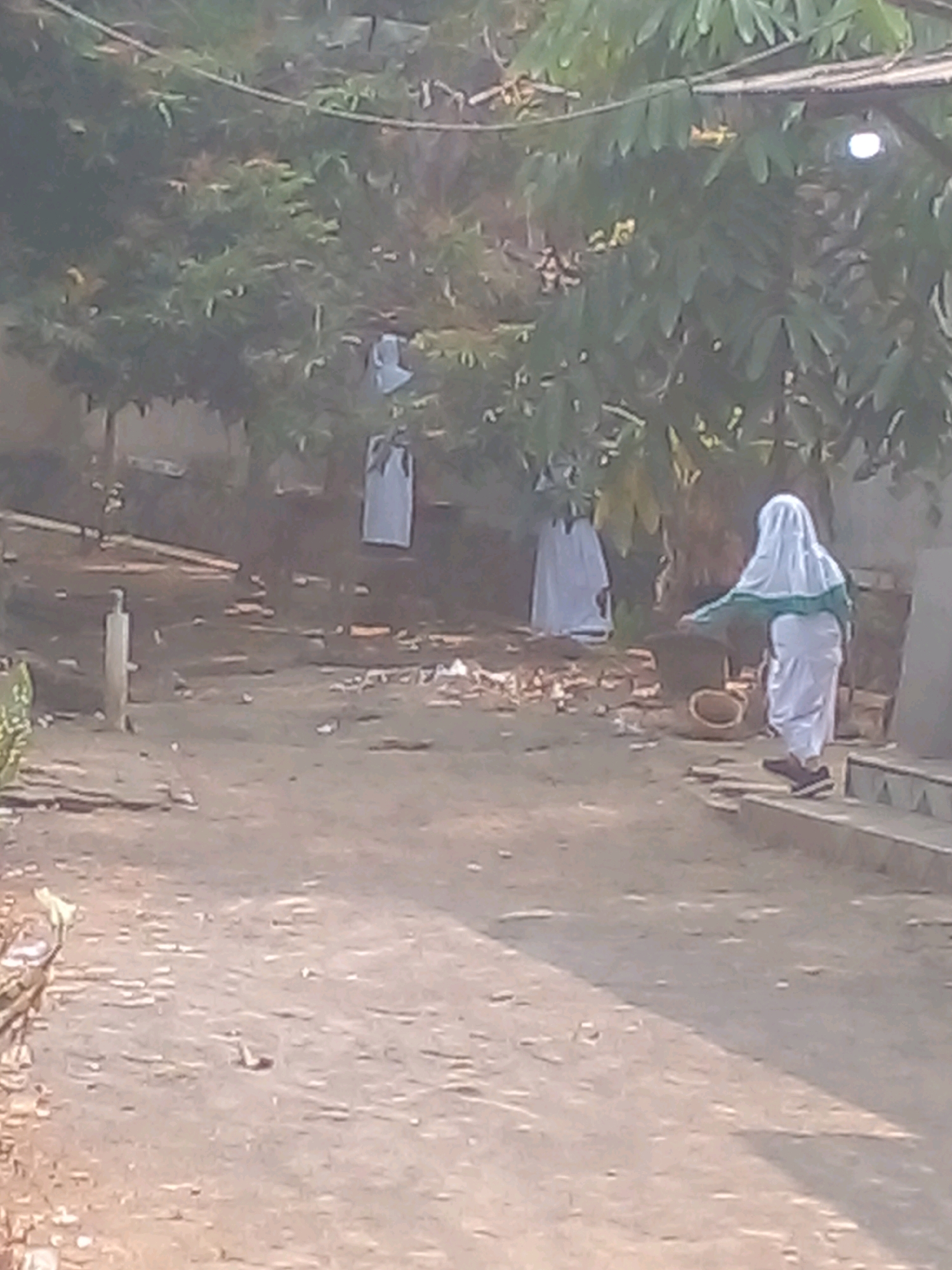In Banyuputih, Indonesia, a scene unfolds in a dirt-covered outdoor area with scattered debris. In the background, partially obscured by trees, two figures in white garments, possibly hijabs, are visible. One is hanging or suspended, while the other appears to be standing. In the foreground, a person wearing a white hijab and green trim, along with dark shoes, is positioned to the right, facing left and bending over a large, dark, circular object with an opening. The environment is natural, with foliage and trees dominating the upper portion of the image, suggesting a rural or semi-rural setting within the city.