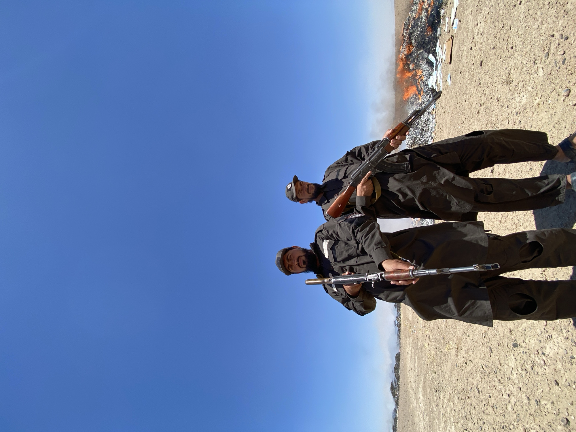 Two men, dressed in dark, loose-fitting clothing and caps, stand in a barren, dusty landscape under a clear blue sky. Each man is holding a rifle. The man on the left, with a beard, holds his rifle vertically in front of him. The man on the right holds his rifle diagonally across his body. Behind them, to the right, is a large pile of burning material, emitting smoke and flames. Scattered on the ground in front of the fire are various items, some of which appear to be paper or cardboard. The ground is dry and sandy, with small stones and debris. The scene appears to be outdoors, in a remote or undeveloped area. The location is identified as the city of Mandisar, Afghanistan.
