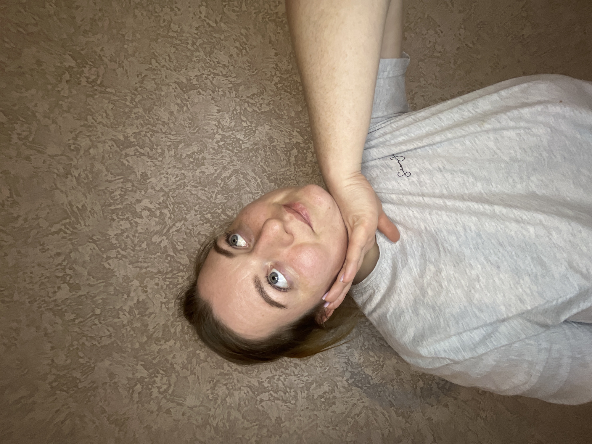 A woman with light skin and blue eyes is lying down on a surface with a mottled brown texture. She is wearing a light grey t-shirt. Her right hand is positioned near her face. The location of the image is Раздолье, Russia.