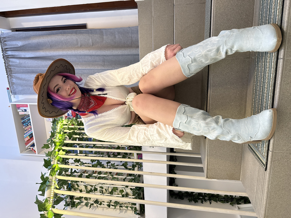 A woman with purple hair wearing a cowboy hat and red bandana, poses in a white blouse and light brown shorts. She's seated on a stairwell with light blue cowboy boots extended. Ivy and string lights decorate the stair railing. Behind her, light grey curtains hang above a white bookshelf filled with books. She smiles at the camera.