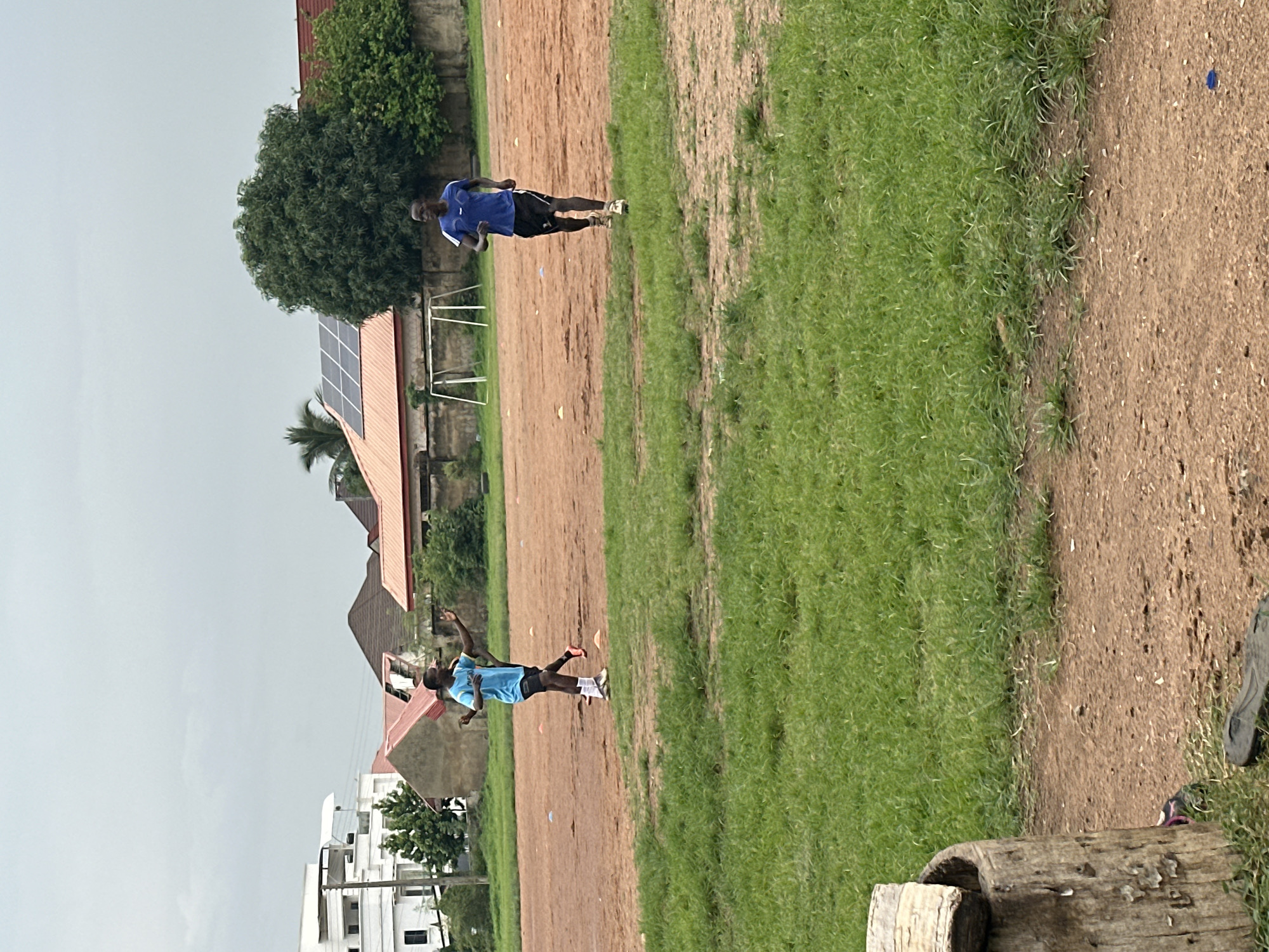 Two young men are seen on a dirt and grass field, likely a sports ground, in Abuja, Nigeria. The man on the right is running in a blue t-shirt and black shorts, facing forward with his body angled slightly to the left. The man on the left, wearing a blue t-shirt and black shorts with white socks, is in motion, appearing to have just thrown or is about to catch an object, with his arms outstretched and body turned. In the background, buildings with orange-tiled roofs and white structures are visible, along with a large green tree and a soccer goal. The sky is overcast.