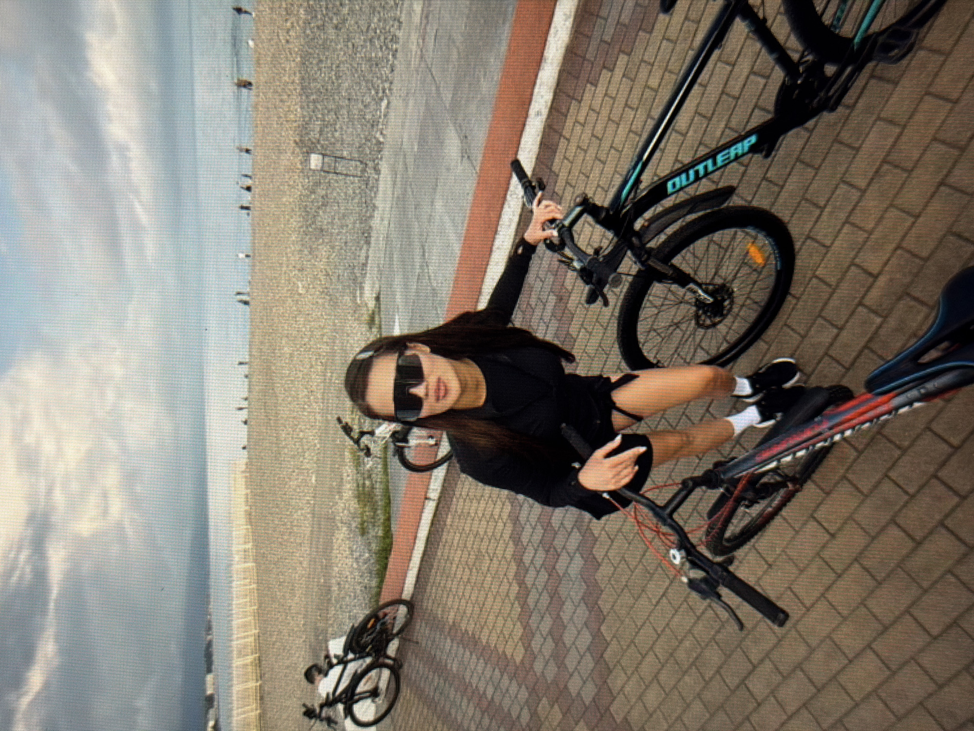 A woman with dark hair and sunglasses poses with two bicycles on a paved promenade overlooking a pebble beach and the sea. The woman is wearing a black outfit and white socks with black shoes. The sky is cloudy. In the background, several people are visible on the beach and in the distance. The bicycles are parked on the promenade. One bicycle is black with teal accents and the brand name "OUTLEPS" visible on the frame. The other bicycle is red and black. The scene appears to be located in Sirius, Russia, given the context.