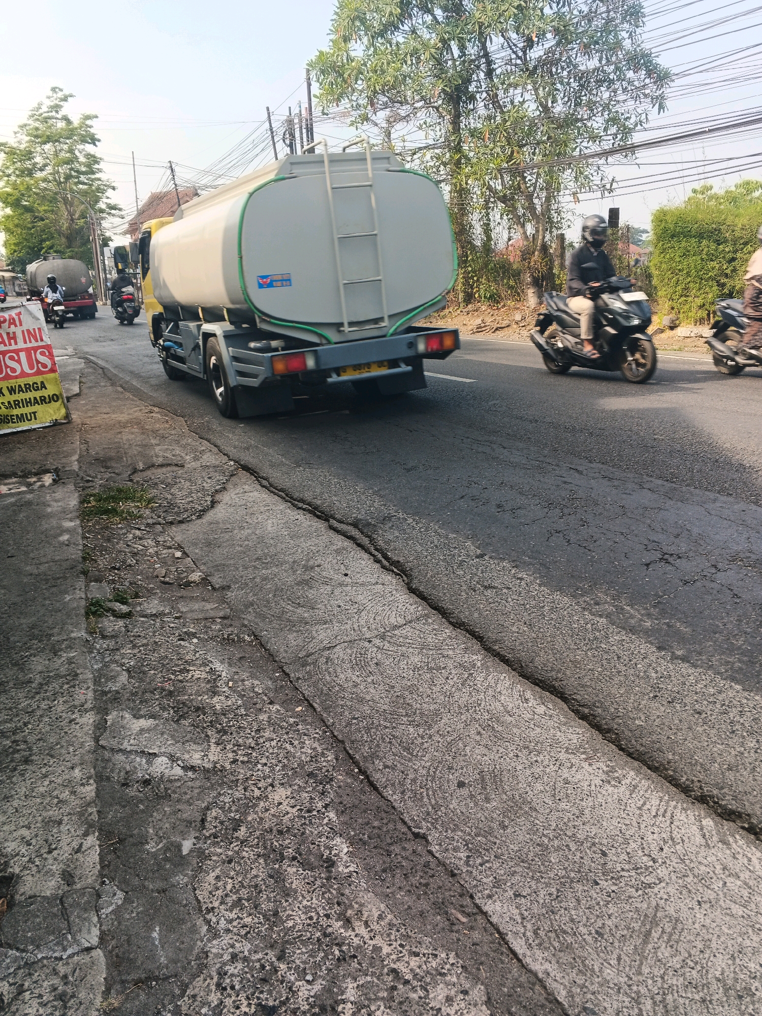 The image captures a street scene in Ungaran, Indonesia, focusing on a yellow water tanker truck in motion. The truck's large, cylindrical tank dominates the right side of the frame. To the left of the truck, a yellow and red sign is partially visible, with Indonesian text reading "PAT INI KHUSUS." Several motorcycles are also present. One motorcycle, ridden by a person wearing a helmet and a dark jacket, is in the foreground to the right of the tanker. Another motorcycle is visible behind the tanker, and a third is further in the background, to the left of the tanker. The road surface appears worn, with visible cracks. The environment includes trees with green foliage lining the street and utility poles with numerous wires overhead, suggesting an urban or semi-urban setting. The sky is clear and bright, indicating daylight. The perspective is from a low angle, looking slightly upwards towards the vehicles.