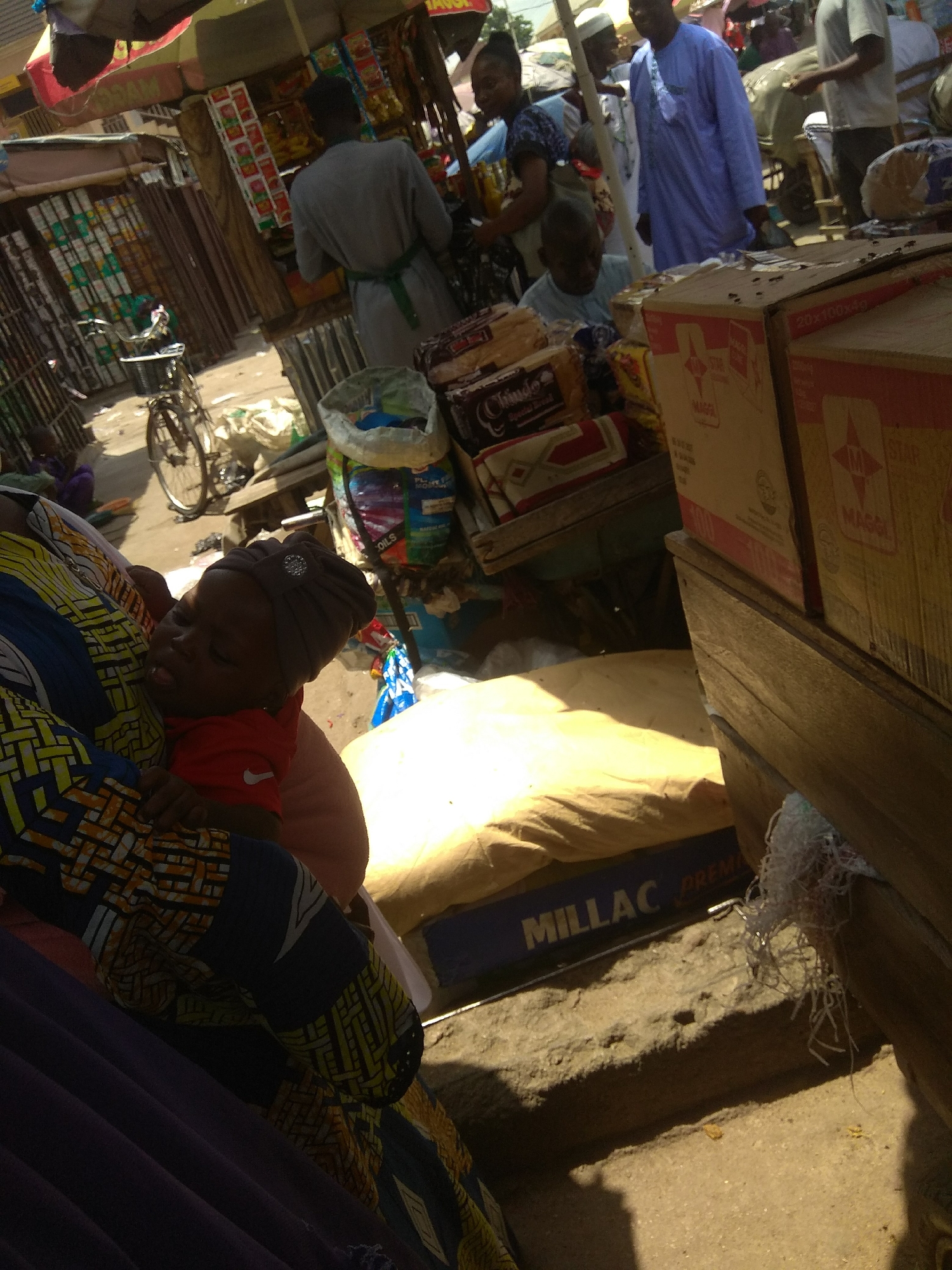 The image depicts a bustling outdoor market scene in Jalingo, Nigeria. In the foreground, a woman is seen carrying a sleeping child. The child, wearing a red shirt and a dark headscarf adorned with a sparkly embellishment, is nestled against the woman's shoulder. The woman is dressed in a vibrantly patterned traditional fabric. Behind them, the market unfolds with stalls laden with various goods, including bags of what appear to be grains or flour, and boxed products.
Several people are visible in the background, engaged in market activities. One man, wearing a blue traditional outfit, stands near a stall. Another man in a grey tunic and a green sash is seen from the back, possibly attending to customers or arranging merchandise. A bicycle is parked nearby, suggesting a mode of transportation for shoppers or vendors. The market appears to be well-stocked, with a variety of items displayed, including packaged foods with labels like "Choco" and "MILLAC PREMIUM."
The lighting suggests it is daytime, likely late morning or early afternoon, with the sun casting shadows. The overall atmosphere appears lively and typical of a busy marketplace. The ground is unpaved and dusty, common in such outdoor settings. The presence of numerous people and diverse goods indicates a lively commercial exchange.