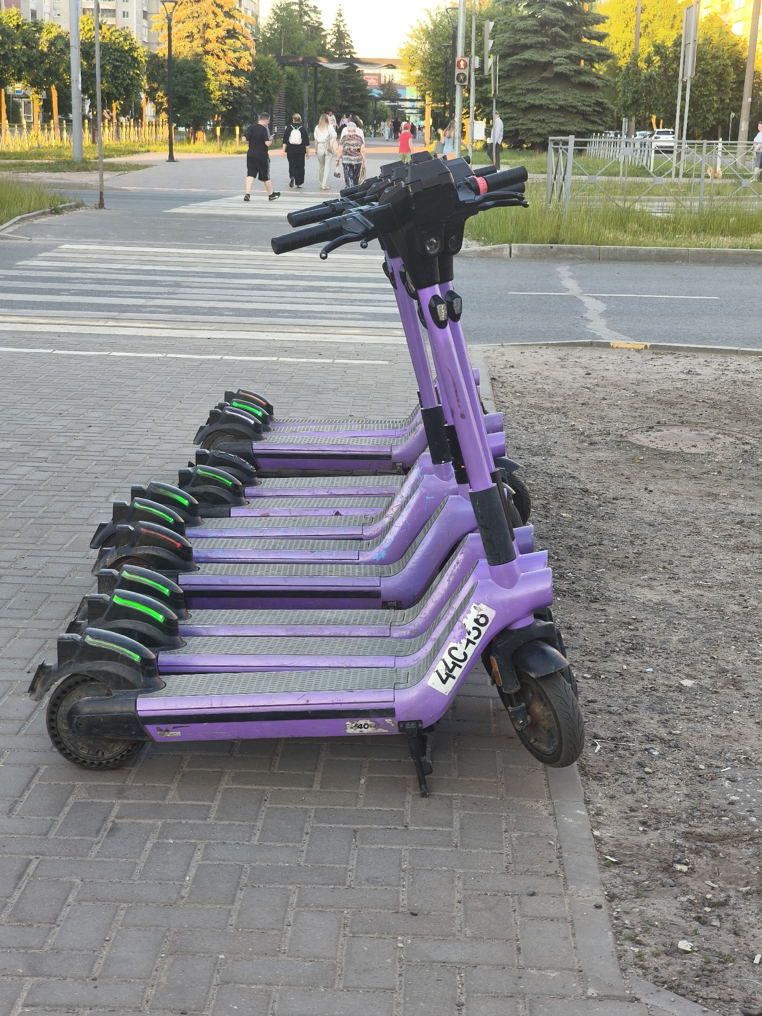 The image depicts a row of parked purple electric scooters on a paved sidewalk next to a crosswalk. In the background, people are walking across the crosswalk. The environment appears to be an urban street with trees and buildings visible. The lighting suggests it is daytime, possibly late afternoon or early evening, with sunlight casting shadows and illuminating the scene. The scooters are lined up neatly, with some displaying green lights on their rear fenders. One scooter has the license plate "440-156" visible. The overall impression is a typical urban scene where shared electric scooters are readily available for use.