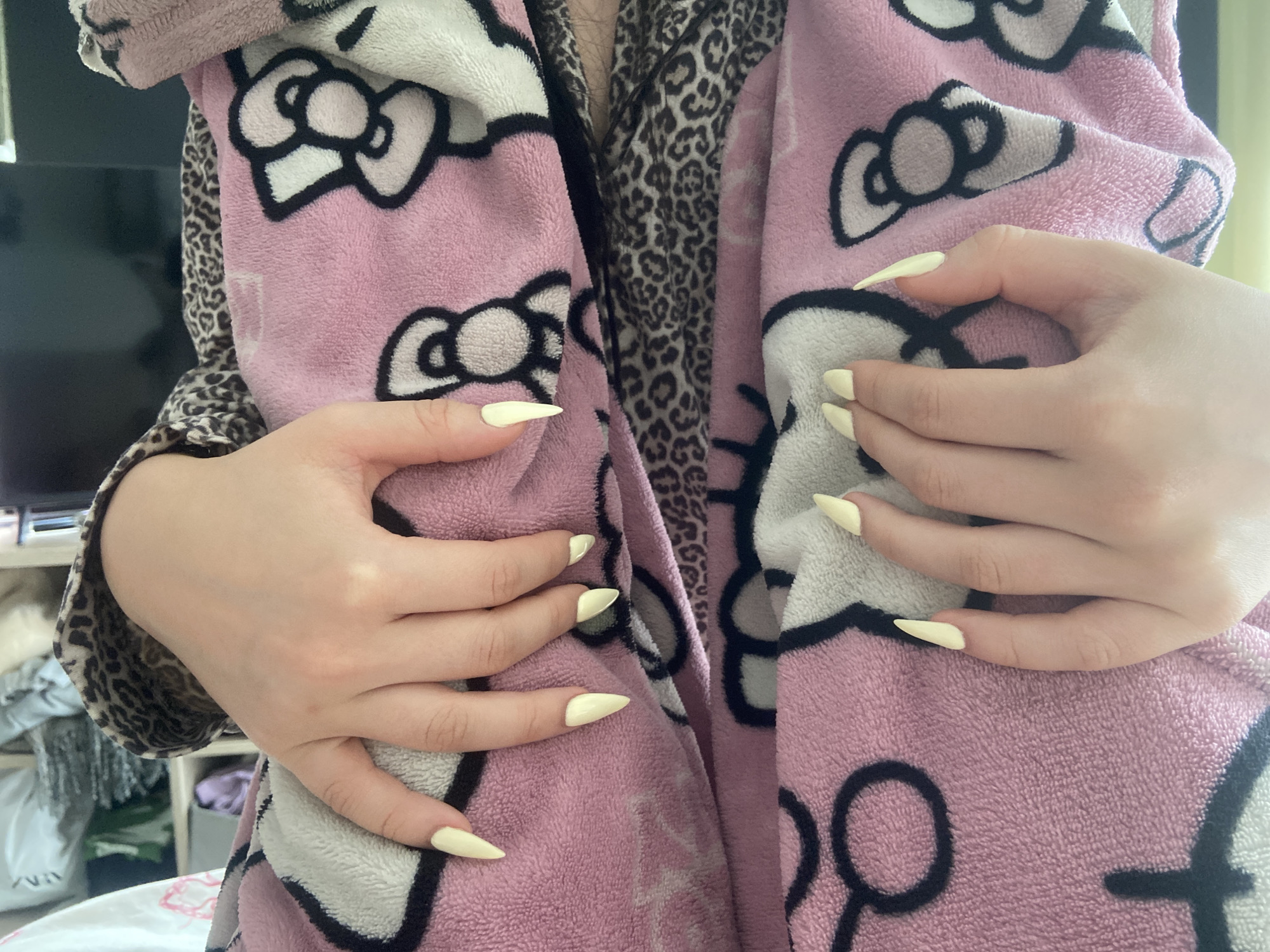 A close-up shot shows a person's hands and torso. The person is wearing a pink and white Hello Kitty patterned robe over a leopard-print garment. The hands are adorned with light yellow stiletto-shaped nails. The background shows a partially visible shelf or dresser with assorted items. The setting appears to be indoors. There is no indication of the location being in London, United Kingdom.
