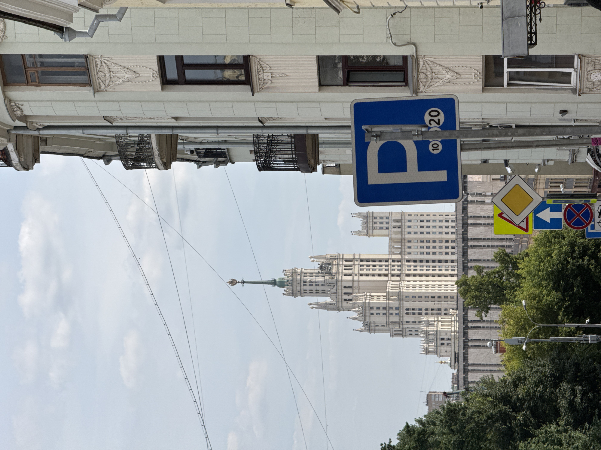 The image depicts a street scene in Moscow, Russia, with a prominent Stalinist skyscraper in the background, likely one of the Seven Sisters buildings. In the foreground, the facade of a lighter-colored building with ornate architectural details and balconies is visible on the right. A blue parking sign with the letter "P" and the numbers "10" and "20" enclosed in circles is mounted on a pole. Several other traffic signs, including a yellow diamond and a blue sign with an upward-pointing arrow, are also present. Lush green trees line the street, partially obscuring other buildings. Wires are strung across the sky above the scene. The sky is partly cloudy.