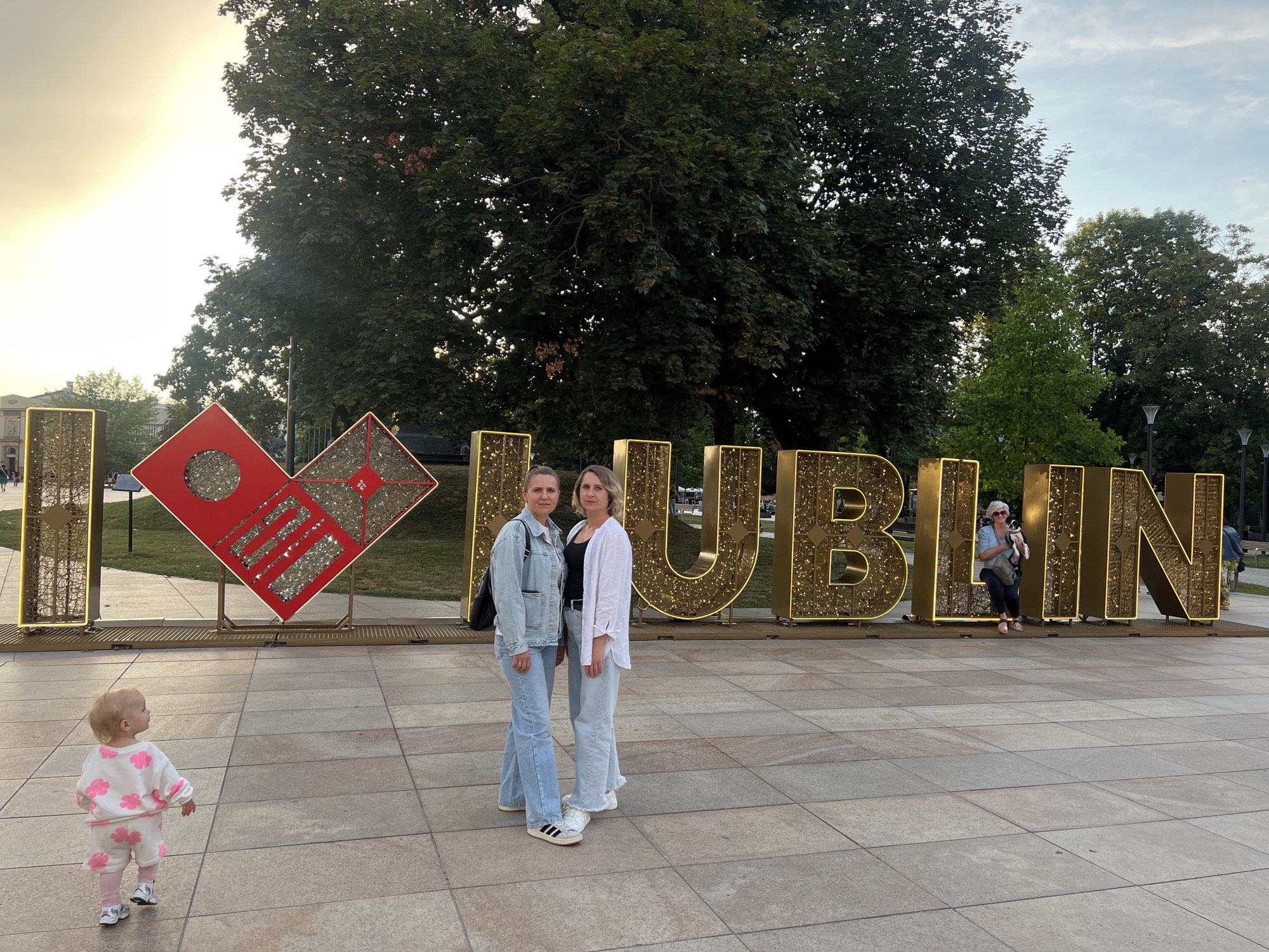 The image depicts an outdoor scene in Lublin, Poland, featuring a large, illuminated sign spelling out "I ♥ LUBLIN". Two women stand in front of the sign, with one on the left wearing a light blue denim jacket and jeans, and the other on the right wearing a white open shirt over a black top and light blue pants. Both women are wearing sneakers. To the left of the sign, a toddler is walking away from the camera, wearing a white outfit with pink floral patterns and pink leggings. Behind the sign, lush green trees form a backdrop, and streetlights are visible further in the background. The ground is paved with large square tiles. The sign itself is gold and illuminated with small lights, and the heart symbol is red and decorated with a pattern. The sky suggests it is either early morning or late afternoon, with a soft, diffused sunlight. A few other people are visible in the distance, further behind the main subjects and the sign.