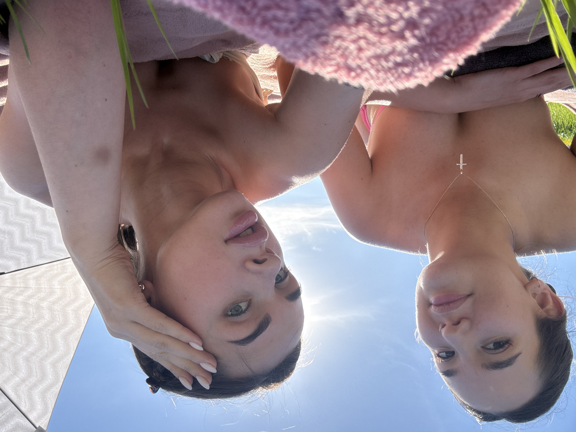 Two women are shown from a low angle, looking down at the camera, against a backdrop of a blue sky and a bit of greenery. The woman on the left has her hand near her face, and the woman on the right is wearing a delicate necklace with a cross pendant. A pink towel or blanket is visible above them. The location is not explicitly identified as Лобаново, Russia, within the image.