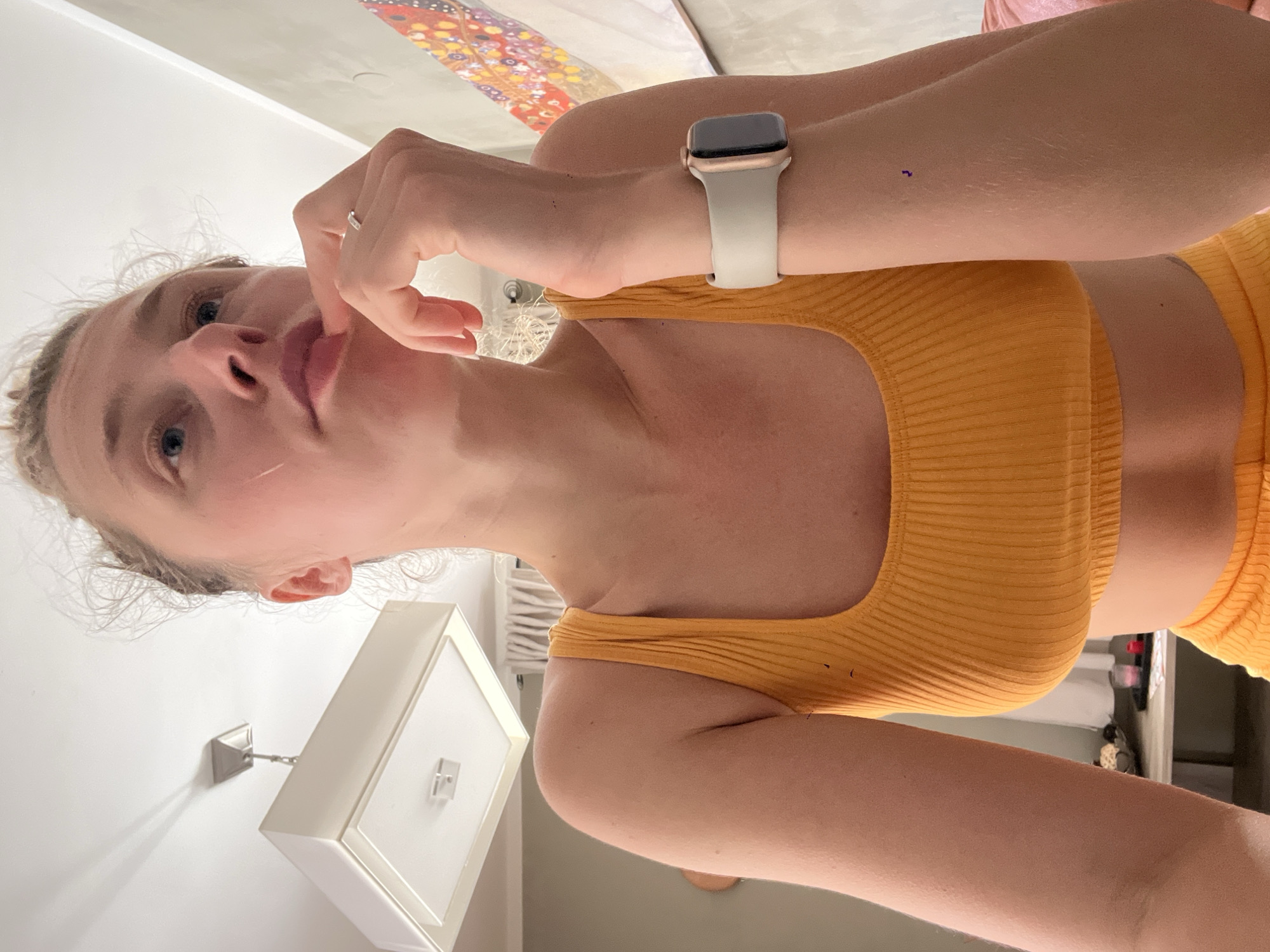 A young woman with fair skin, blonde hair, and blue eyes is holding a finger to her lips. She's wearing an orange ribbed crop top and an Apple Watch with a white band. The setting is indoors, with a white wall and a square, white ceiling light fixture visible. A decorative overhead element with colorful patterns is partially visible in the upper portion of the frame. The location is not identifiable as Bečići, Montenegro, based on the provided image.