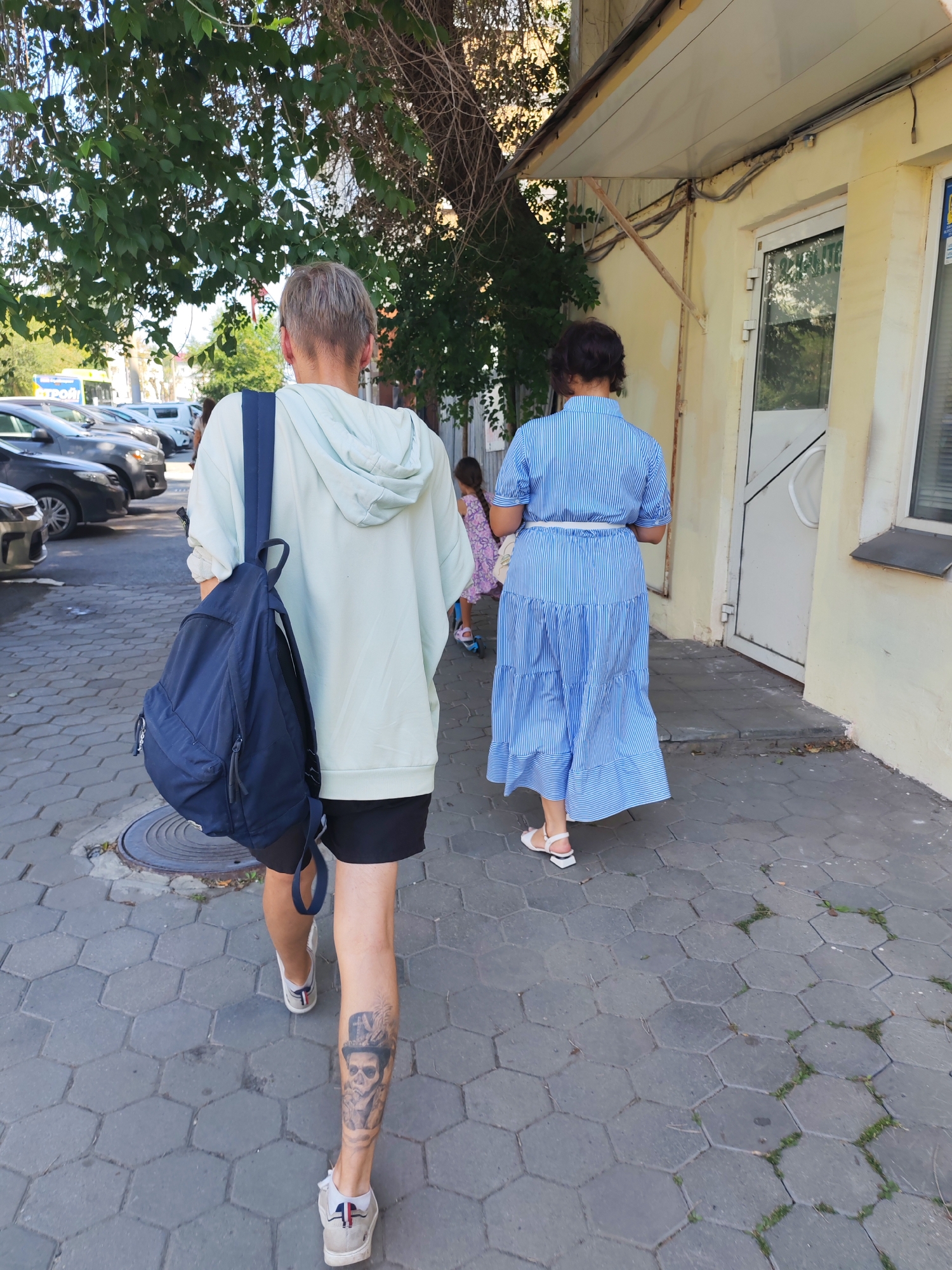 The image shows two people walking away from the camera on a hexagonal-tiled sidewalk in front of a yellow building. The person in the foreground on the left is wearing a light blue hoodie, black shorts, and white sneakers, with a dark blue backpack slung over their left shoulder. They have short blonde hair and a tattoo of a skull wearing a hat on their right calf. To the right of this person, a woman in a blue and white striped dress with white sandals is walking. Further in the background, a child in a patterned dress is visible. Cars are parked along the street on the left side of the image. Overhead, tree branches with green leaves hang above the sidewalk. The building on the right has a white door and a window.