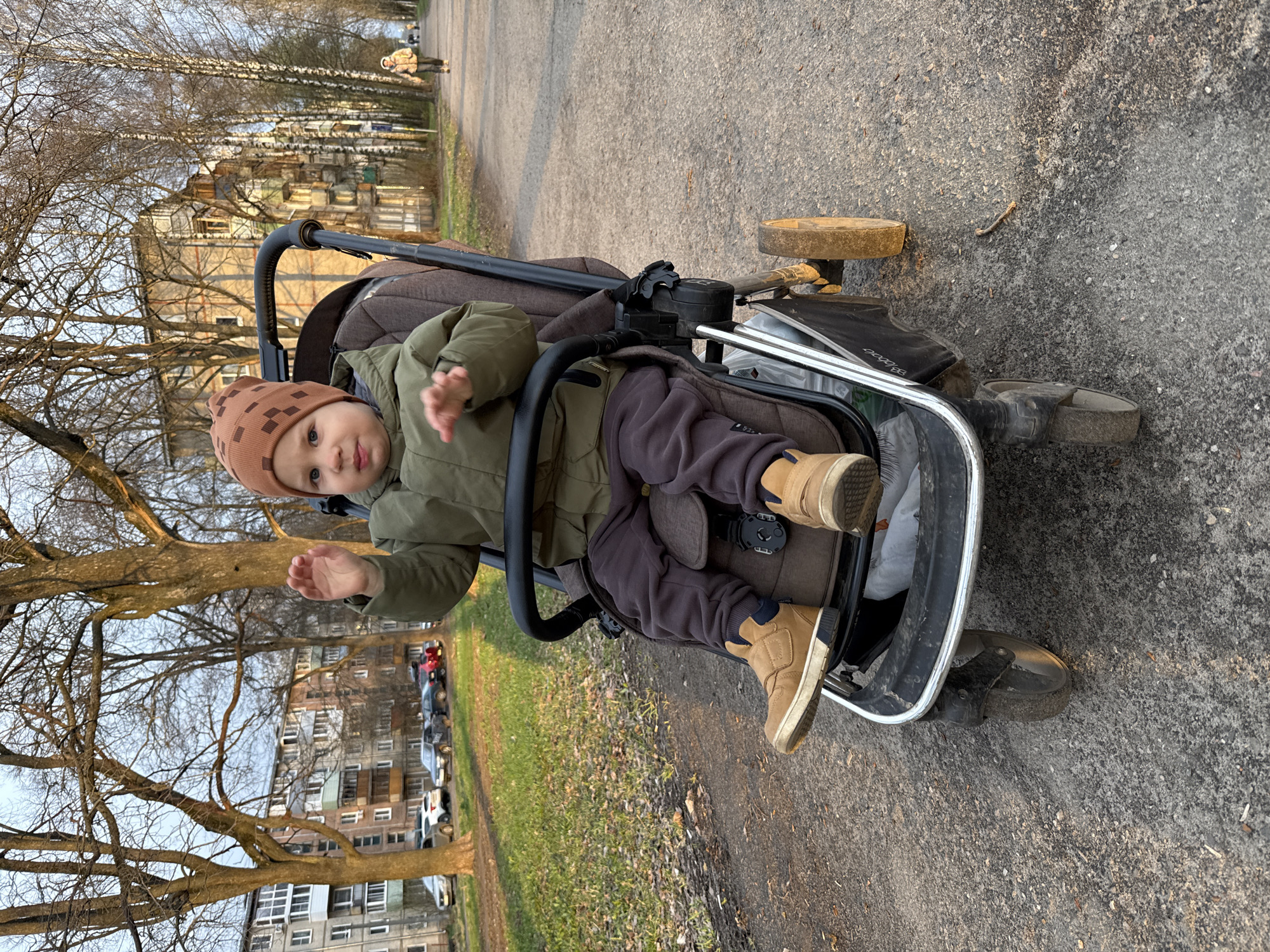 The image shows a young child, likely a toddler, seated in a stroller on an outdoor asphalt path in Rusanovo, Russia. The child is dressed in a green jacket, dark pants, and tan shoes, and wears a brown knit hat with a subtle geometric pattern. Looking directly at the camera with a curious or playful expression, the child has both hands raised. The stroller is brown and grey, with the brand name "UPPABaby" visible on its storage basket and "BB" on a grey component of the frame.
The background features a row of tall, bare-branched trees lining the path, suggesting late autumn, winter, or early spring. Beyond the trees, multi-story residential apartment buildings are visible. In the far distance along the path, another person can be seen walking. The lighting indicates either a late afternoon or early morning "golden hour," casting a warm glow over the scene and suggesting clear weather. The ground adjacent to the path is covered with sparse, dry grass. The scene depicts a quiet, everyday outdoor activity.